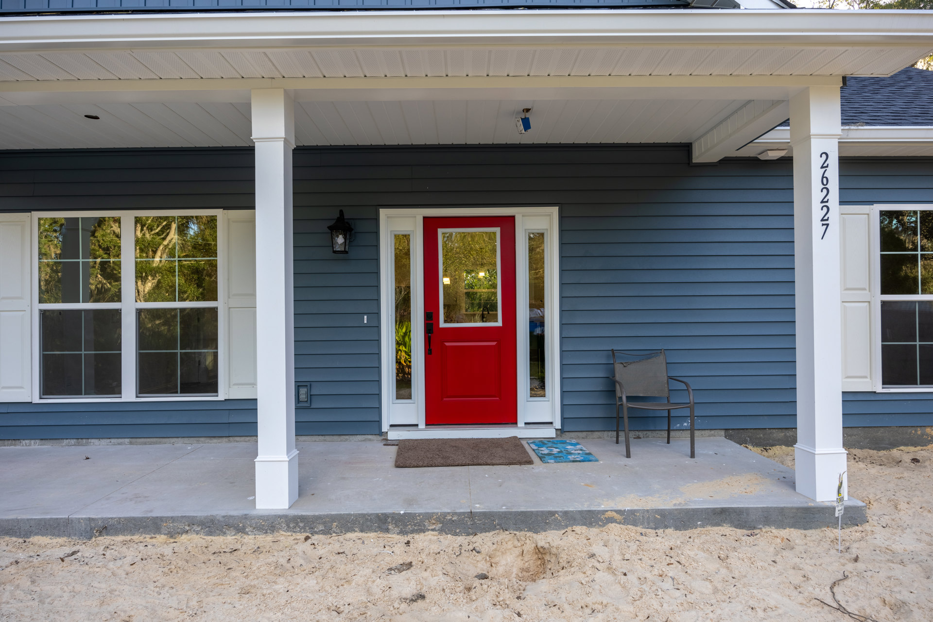 Red front door with glass window, weathered wooden chair with broken seat on covered porch, white siding, sash window reflecting tree branches, metal light fixture above door.