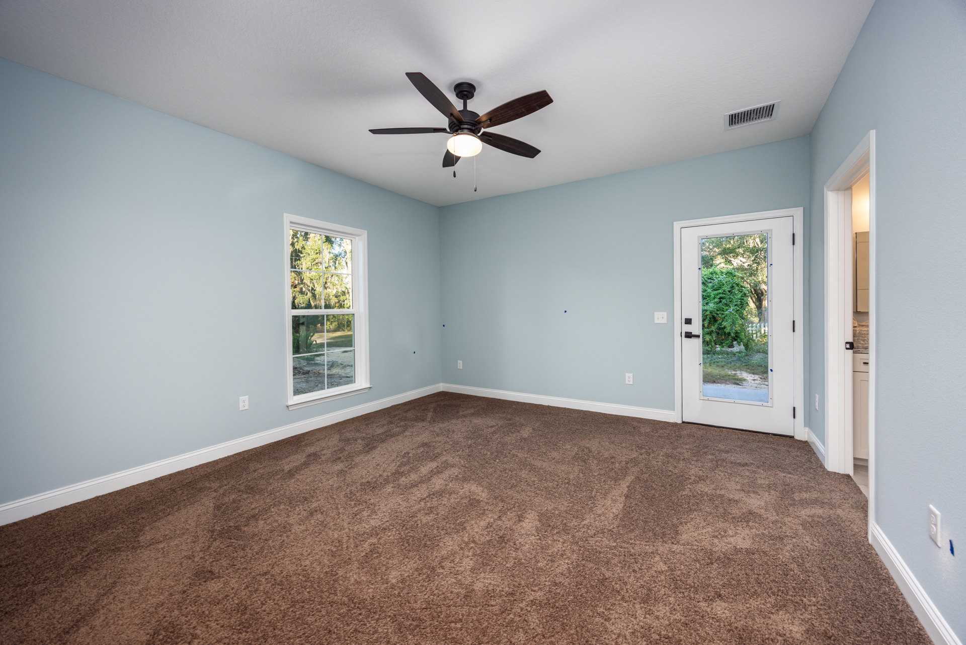 Ceiling fan with light fixture, white plaster ceiling with vent, brown carpet flooring, door opening to garden view, window overlooking trees and grass