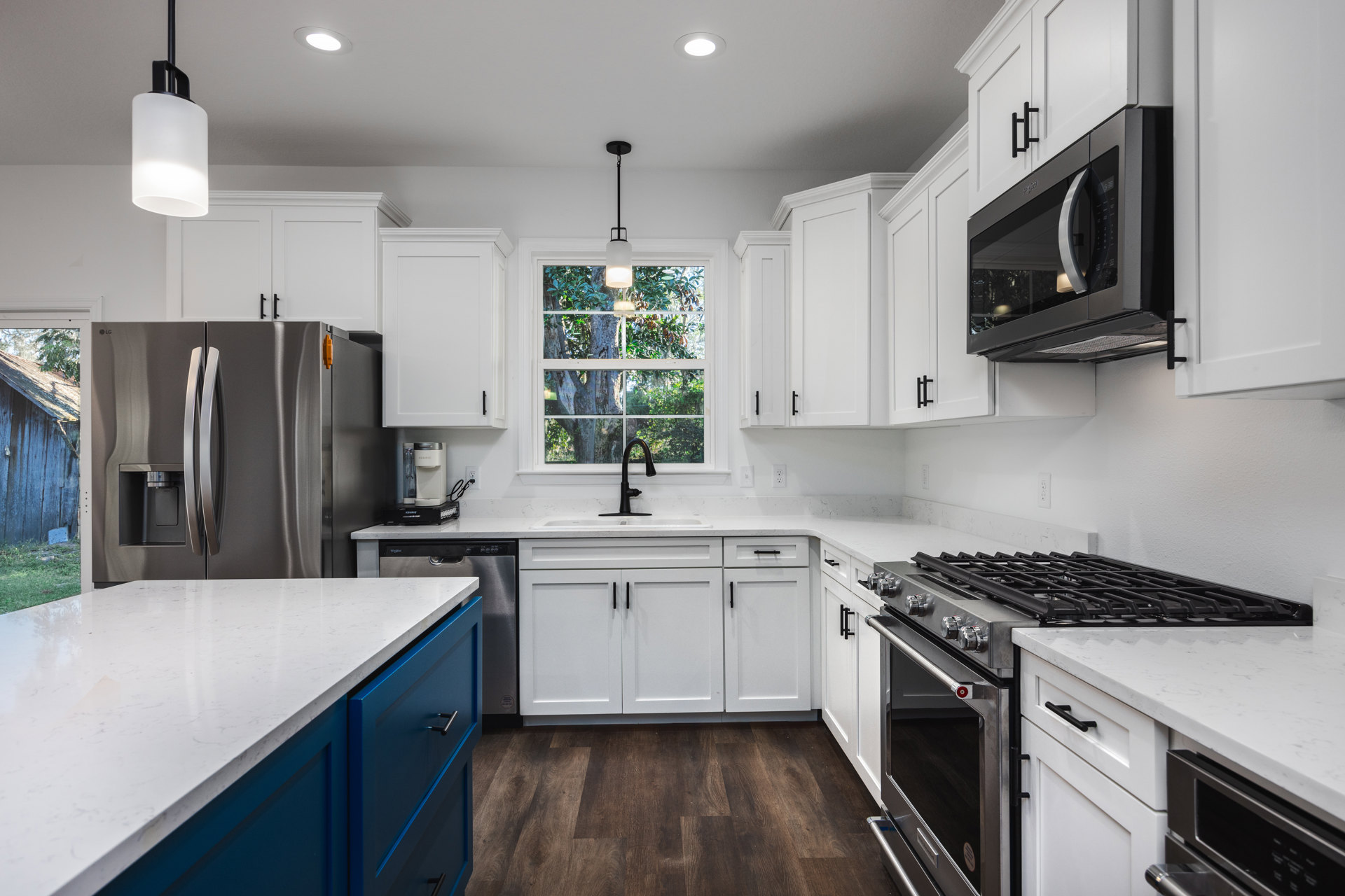 White kitchen cabinets paired with blue lower drawers, white countertop, stainless steel stove and oven, built-in microwave, refrigerator, sink, and window overlooking trees.