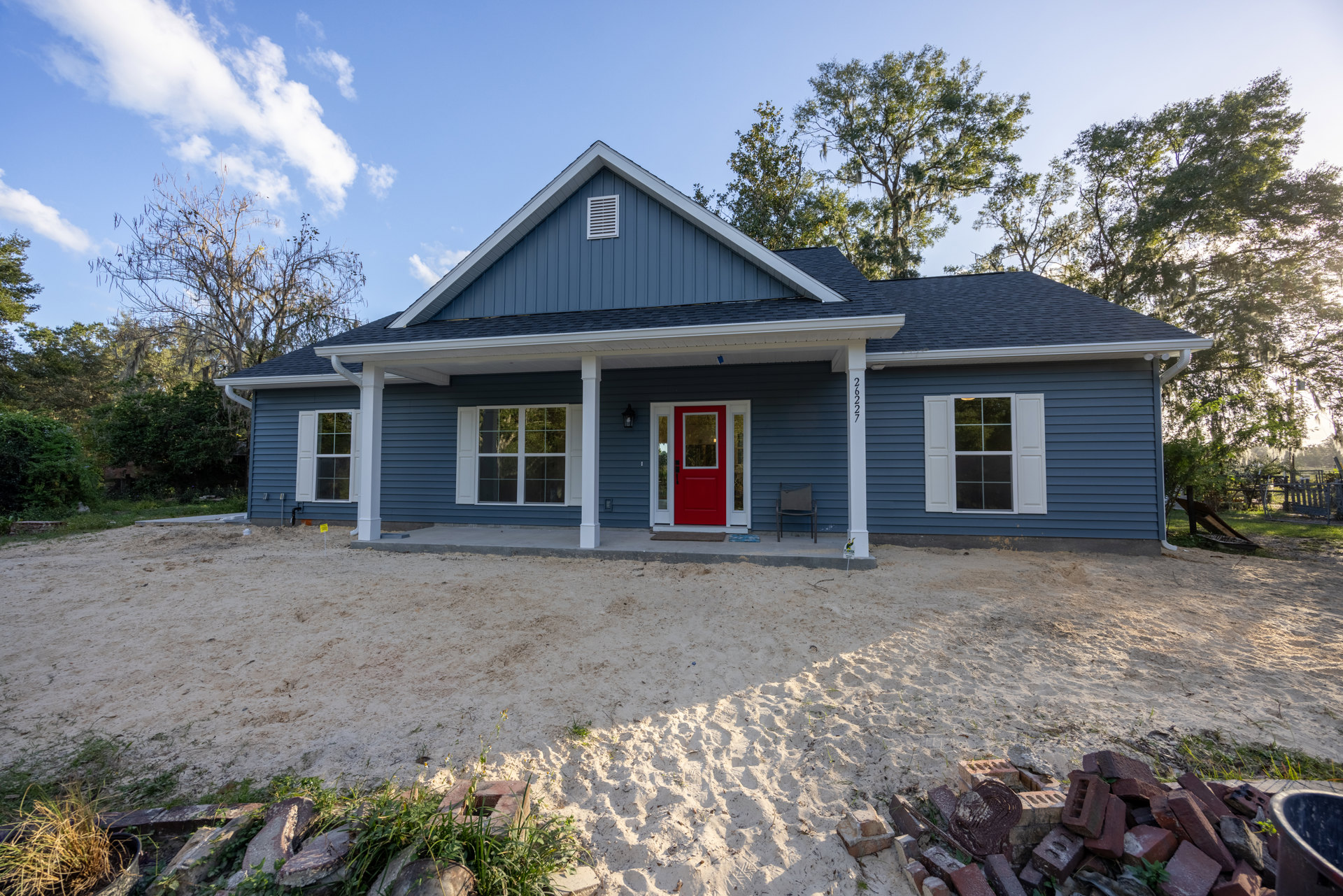 Blue siding house with red front door, white-framed windows with shutters, outdoor chair on porch, sandy yard, small white house visible in background, trees and cloudy sky
