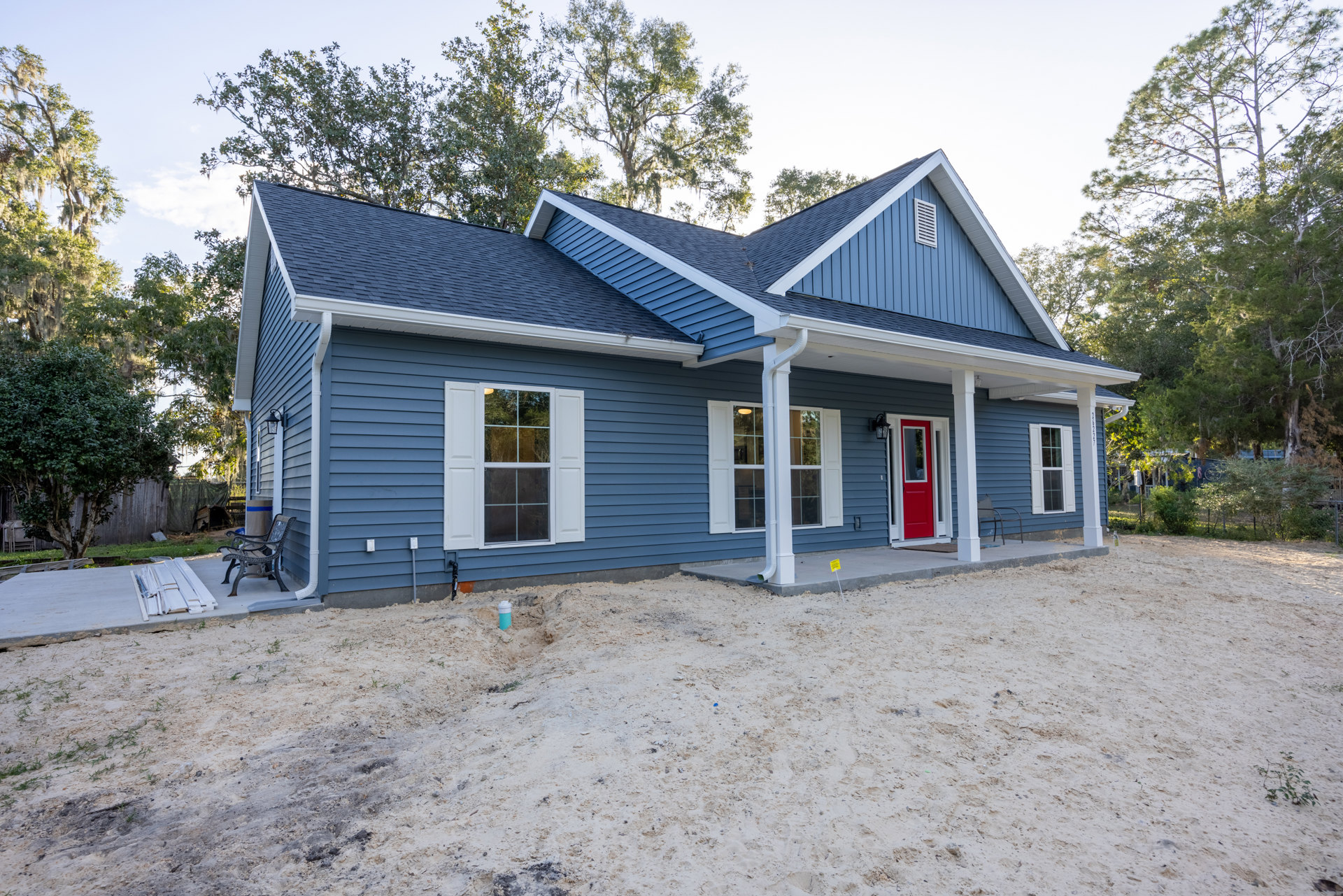 Blue house with white siding, red door framed in white, porch bench, window with white shutters, tree and sky in background, hole in ground near foundation