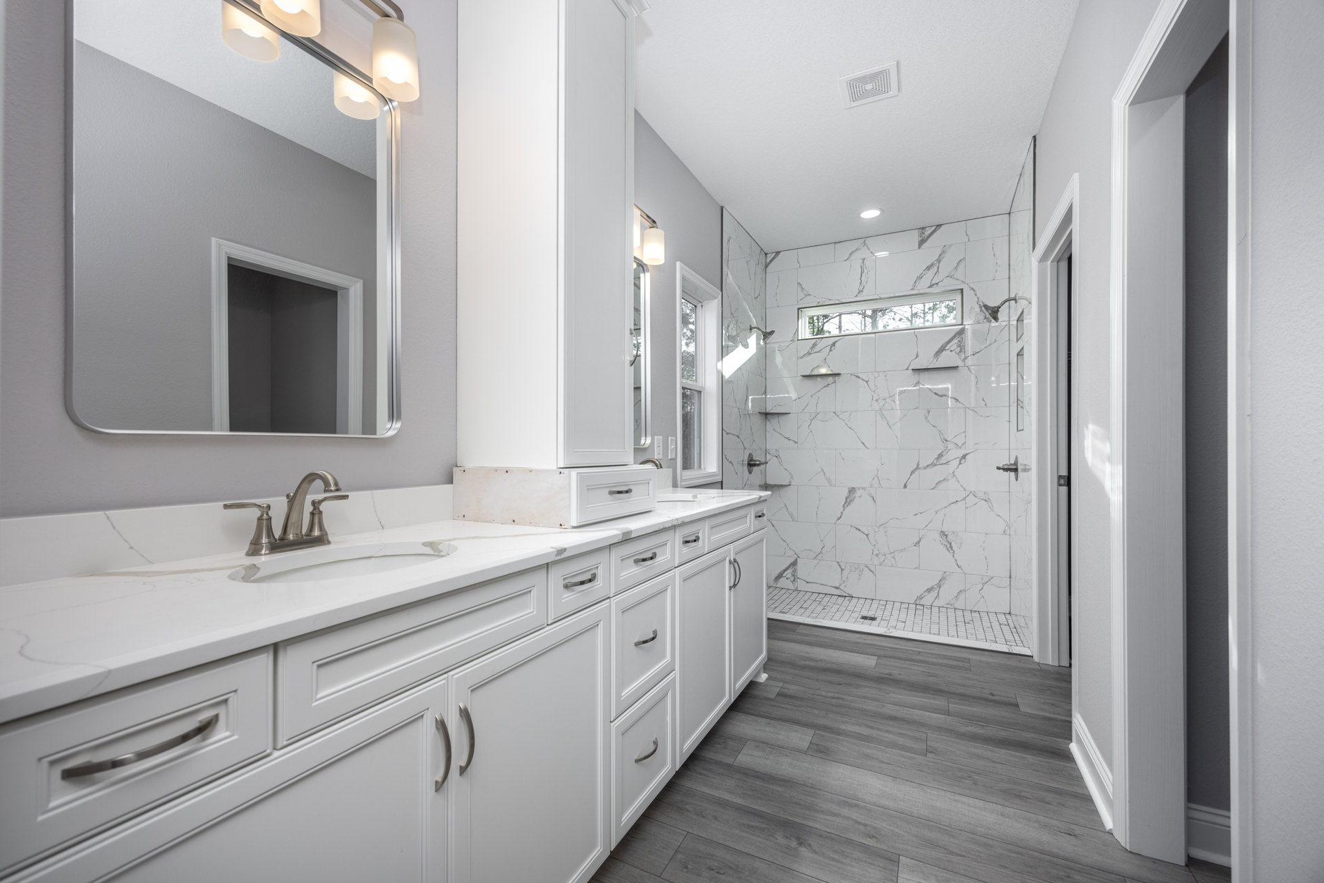 White bathroom with tile shower, white cabinets, quartz countertop, wall-mounted mirror with lights, chrome faucet, and grey painted walls.