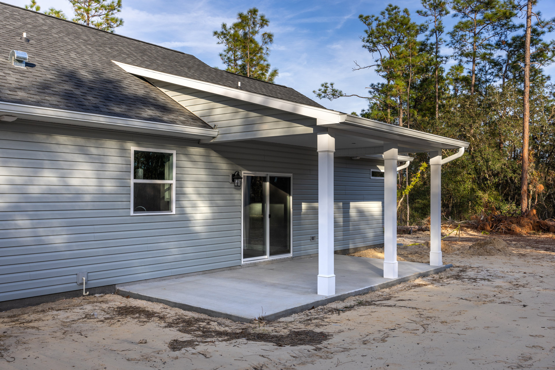 Two-story home with covered front porch, glass-paneled door, illuminated window, metal roof vent, tree branches over roof, concrete patio with scattered sticks, cloudy sky in