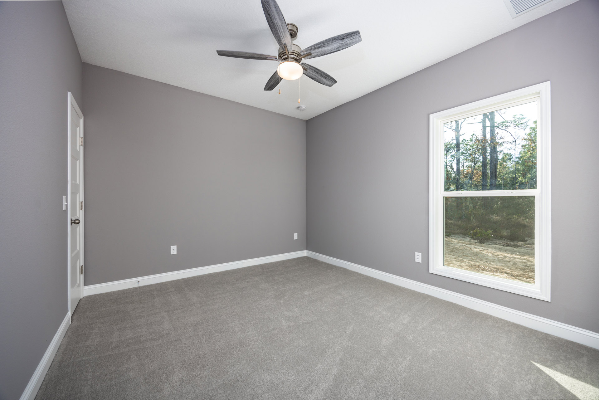 Carpeted room with white trim, ceiling fan with light fixture, large window overlooking trees and dirt path
