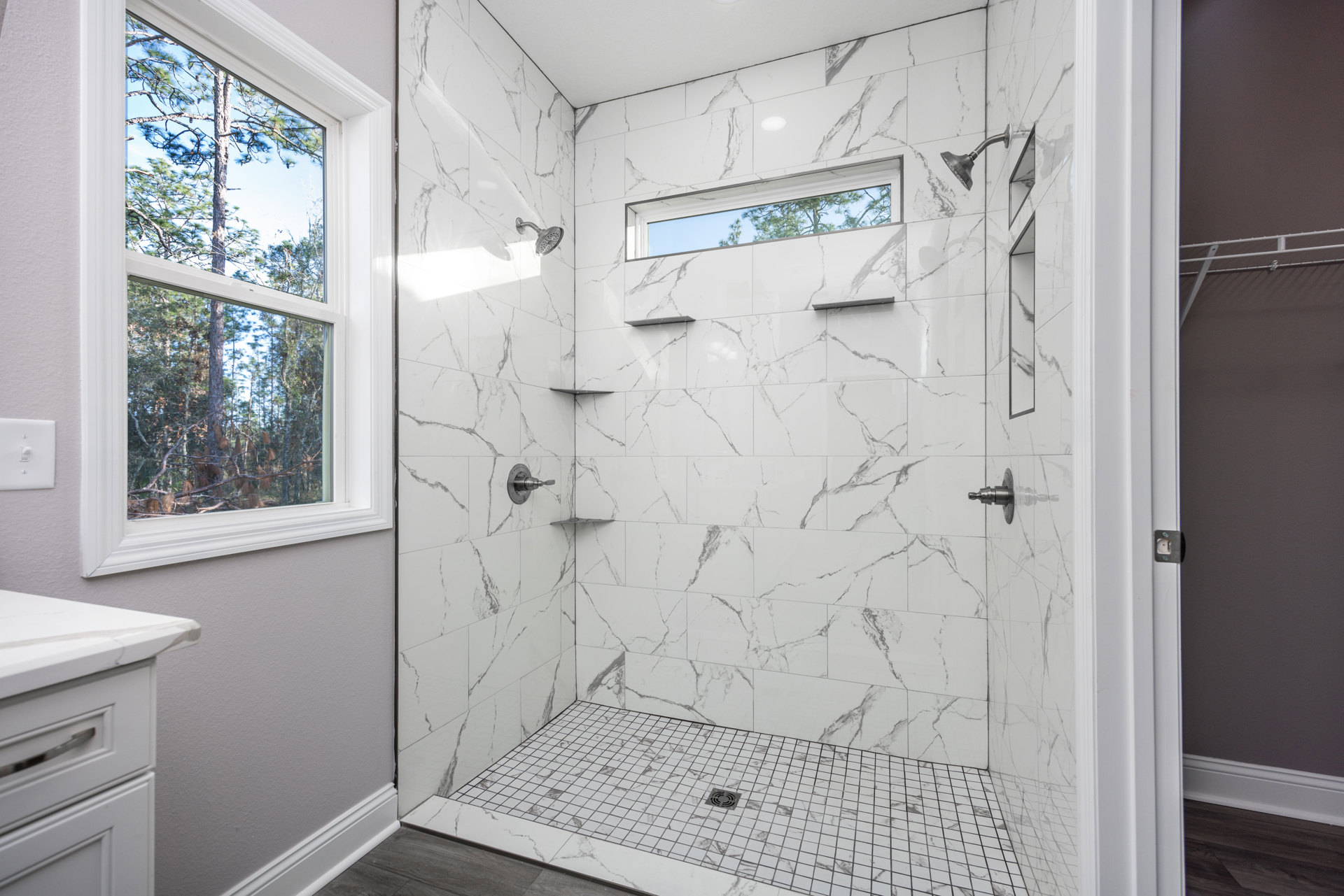 Shower with marble tile walls and floor, window overlooking trees, white countertop with silver handle, close-up of shower knob and drain