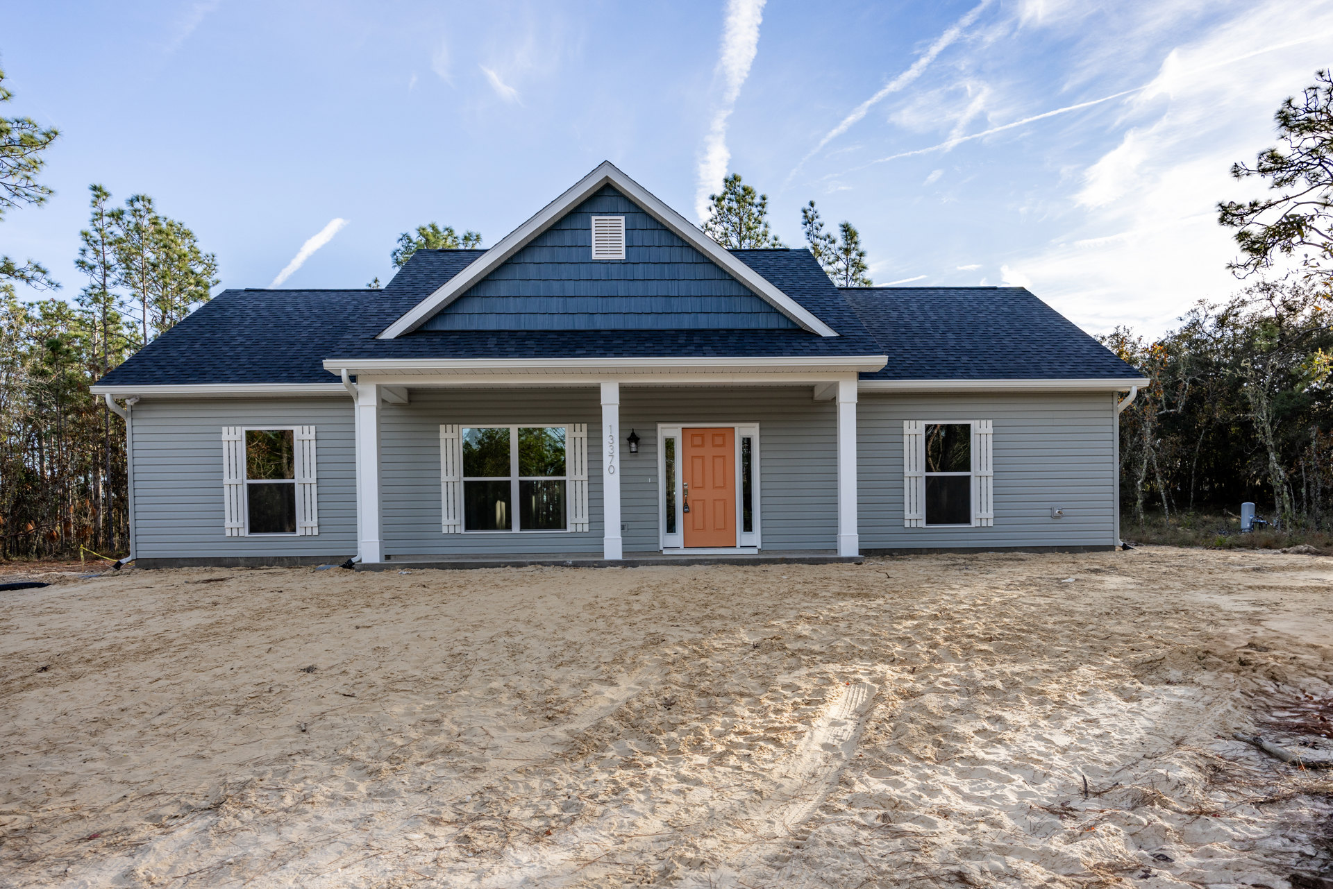 Orange front door and white shuttered window on a custom home, sandy dirt driveway with tire tracks, trees reflected in glass, cloudy sky overhead