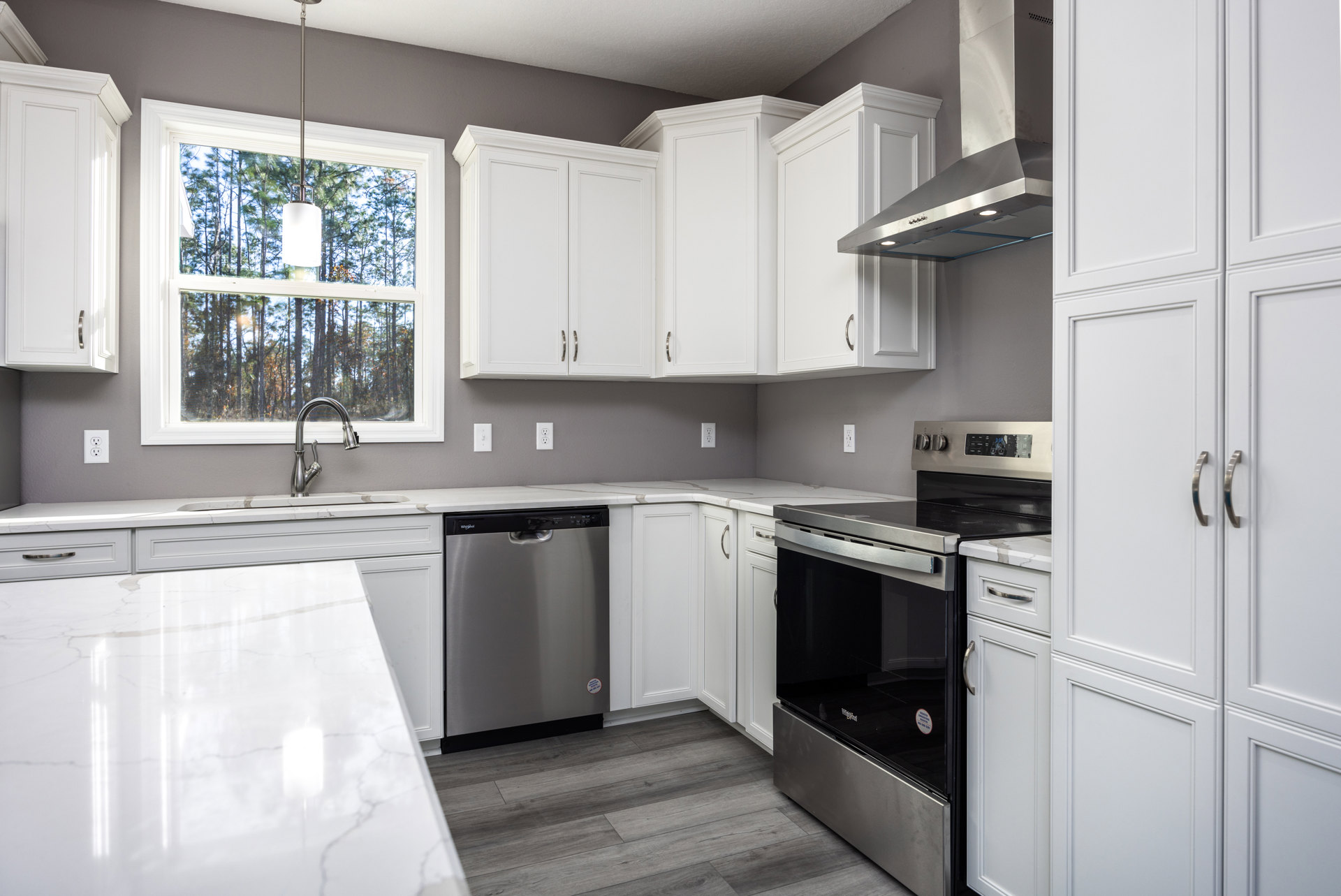 White kitchen with shaker cabinets, stainless steel stove with glass door, built-in dishwasher, white marble floor, white walls, window letting in natural light, black countertop.