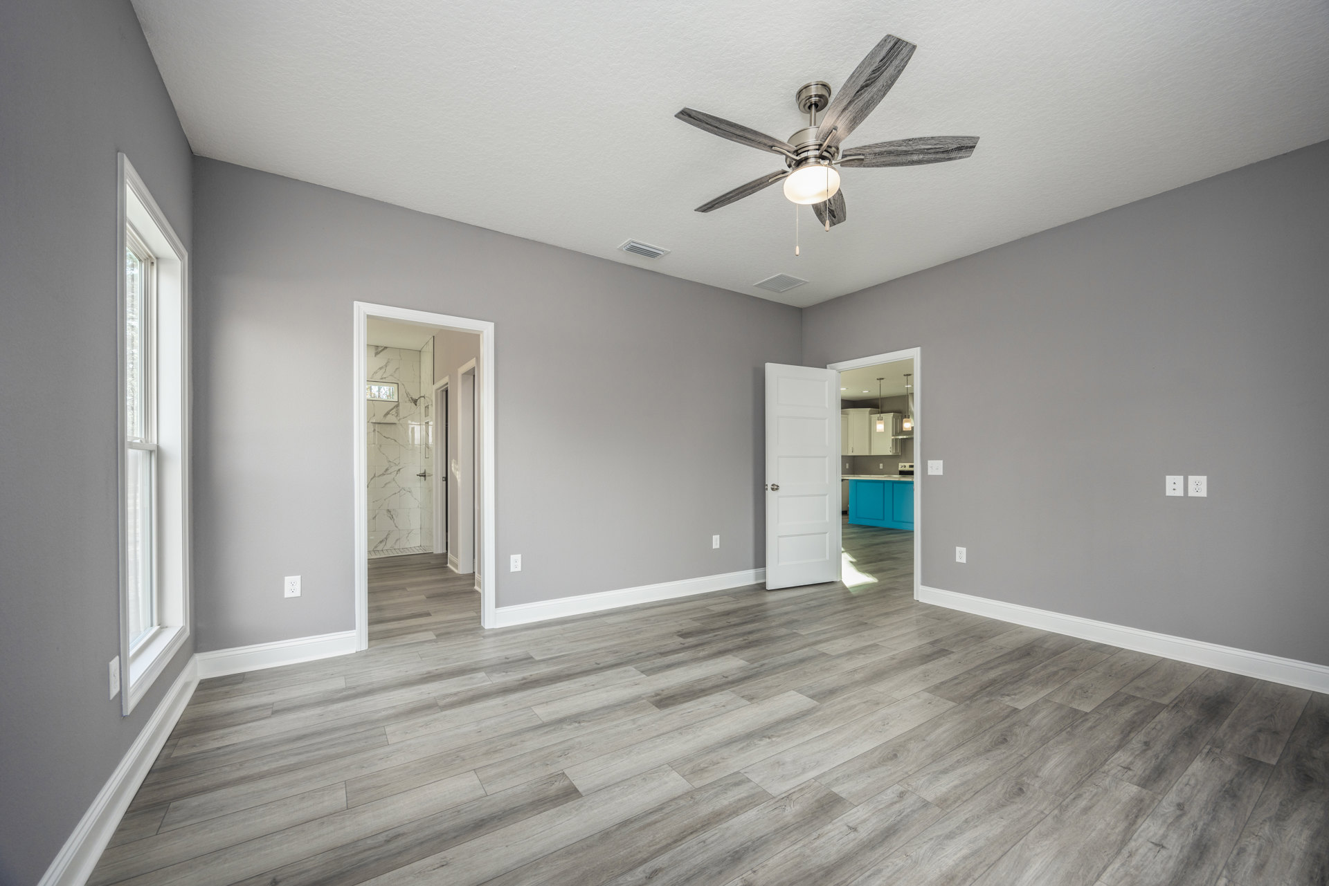 Ceiling fan with light fixture, wood laminate flooring, white paneled door with silver knob, window with white trim, neutral painted walls, partial view of shower with window