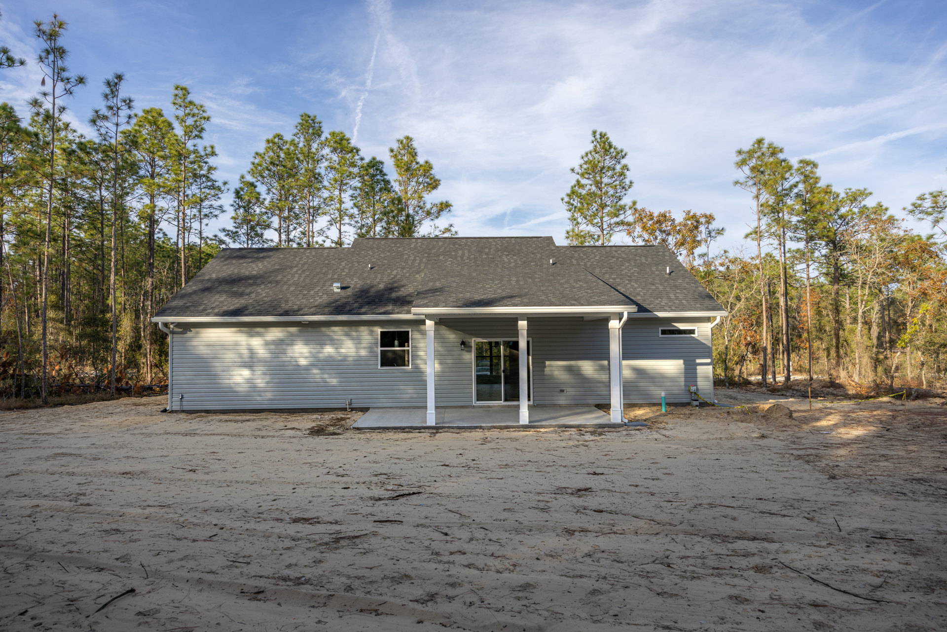 Single-story cottage with wood siding, covered porch, glass door, illuminated window, dirt driveway, and surrounding trees under cloudy sky