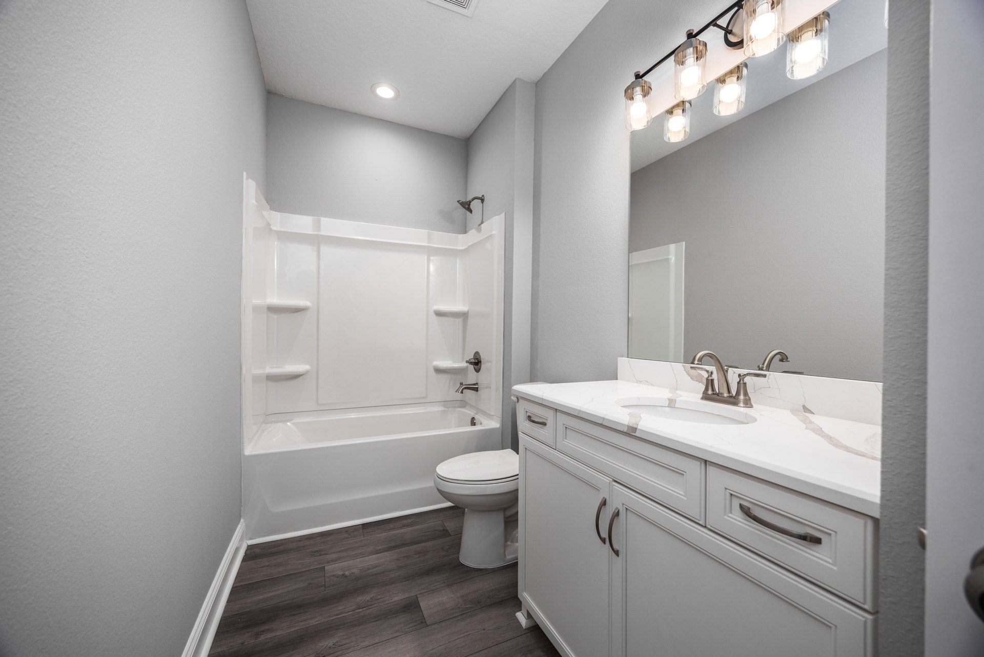 Bathroom with white bathtub, modern sink vanity, closed toilet, wood flooring, white trim, and wall-mounted light fixture with clear glass shades