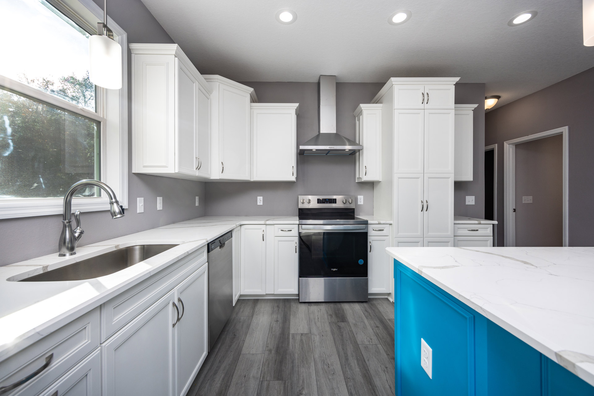 White kitchen cabinets, blue island with white marble countertop, stainless steel oven with black door, ceiling light fixture, sink and faucet beneath window overlooking trees