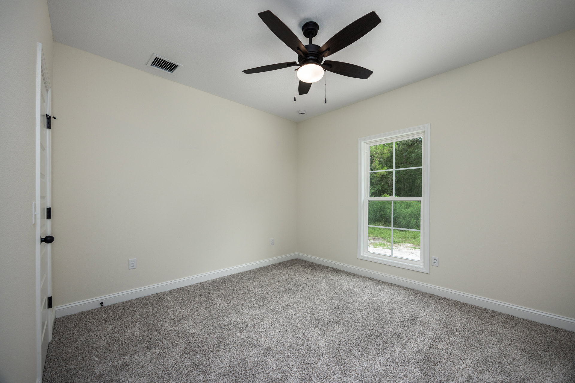 Ceiling fan with integrated light fixture, white plaster ceiling with vent, carpeted floor, large window overlooking trees and grass