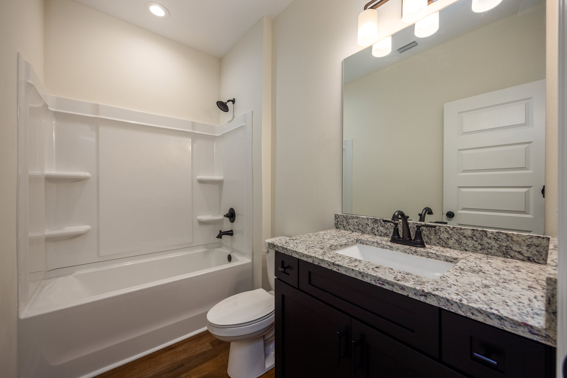 White bathroom with tile floor, white toilet, rectangular sink on stone countertop, wall-mounted mirror above sink, white bathtub adjacent to toilet, white door with black handle