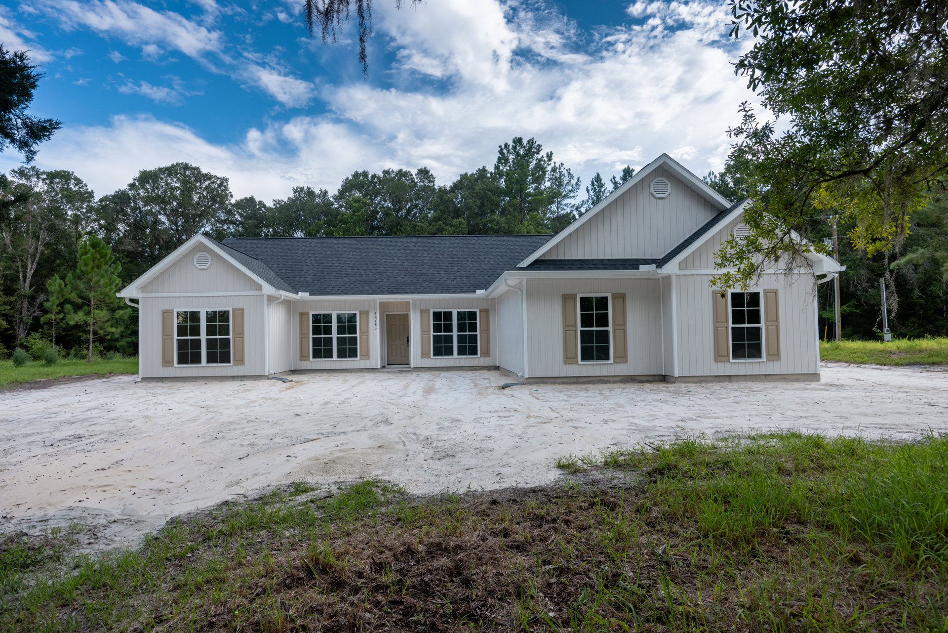 White siding house with white-trimmed windows, paved driveway, dirt landscaping area, and mature trees in the background