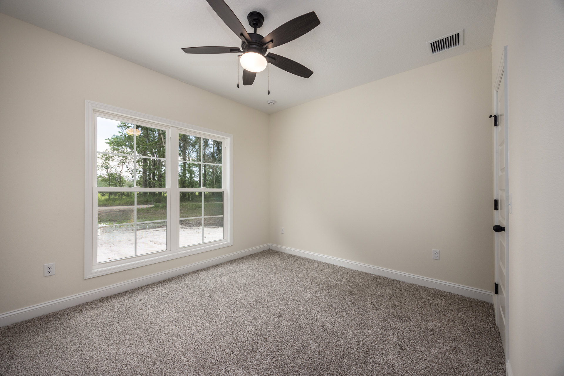 Neutral-toned carpeted room with white walls, ceiling fan with light fixture, large window showing green trees outside, wall vent visible near floor