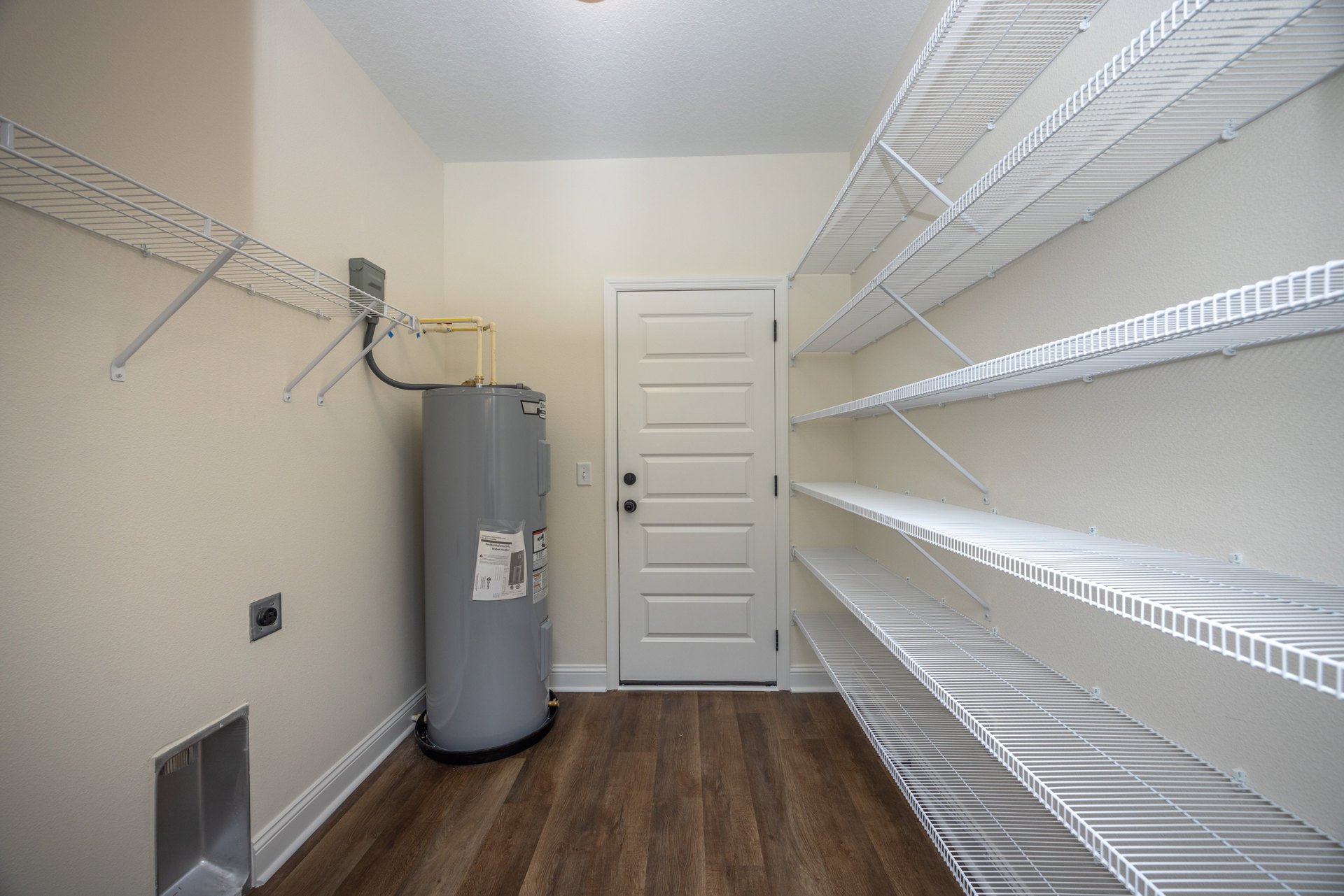 Utility room with white shelves mounted on the wall, large cylindrical metal water heater, white door with black knobs, light-colored plaster walls, and neutral flooring.