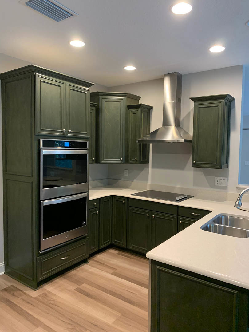 Green kitchen cabinets with brass hardware, stainless steel stove, white quartz countertops, farmhouse sink, light wood flooring, and subway tile backsplash