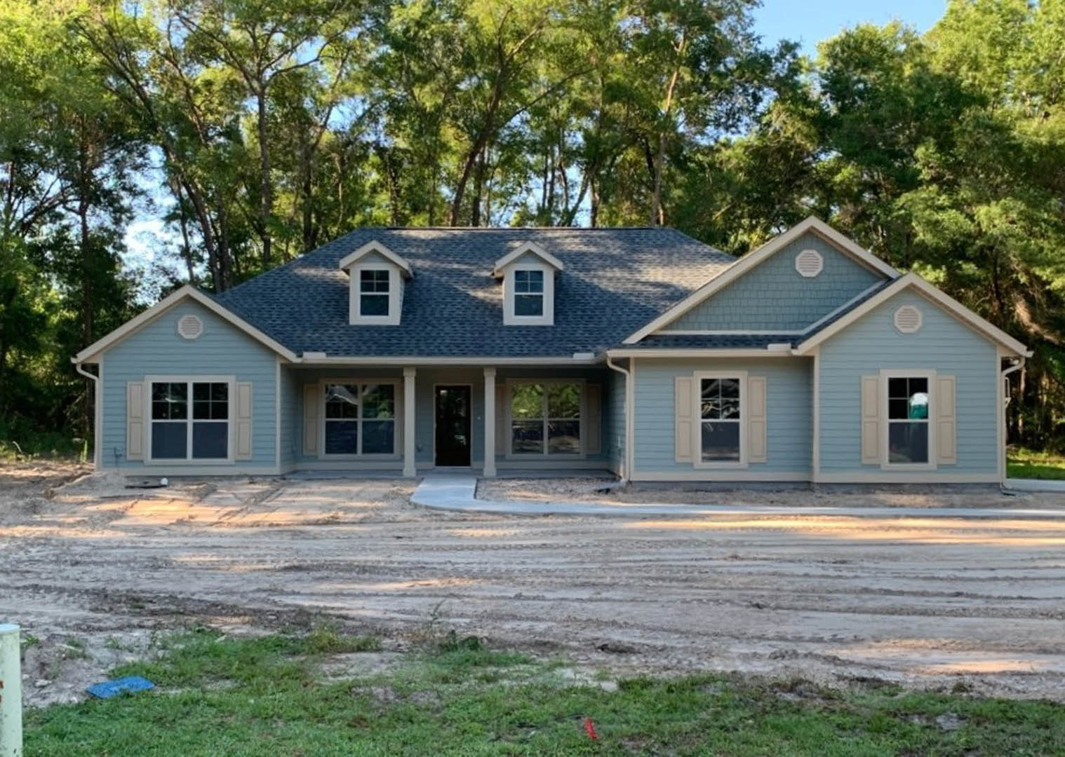 Framed house under construction with blue roof, exposed plywood siding, white vent, and surrounding trees in the background