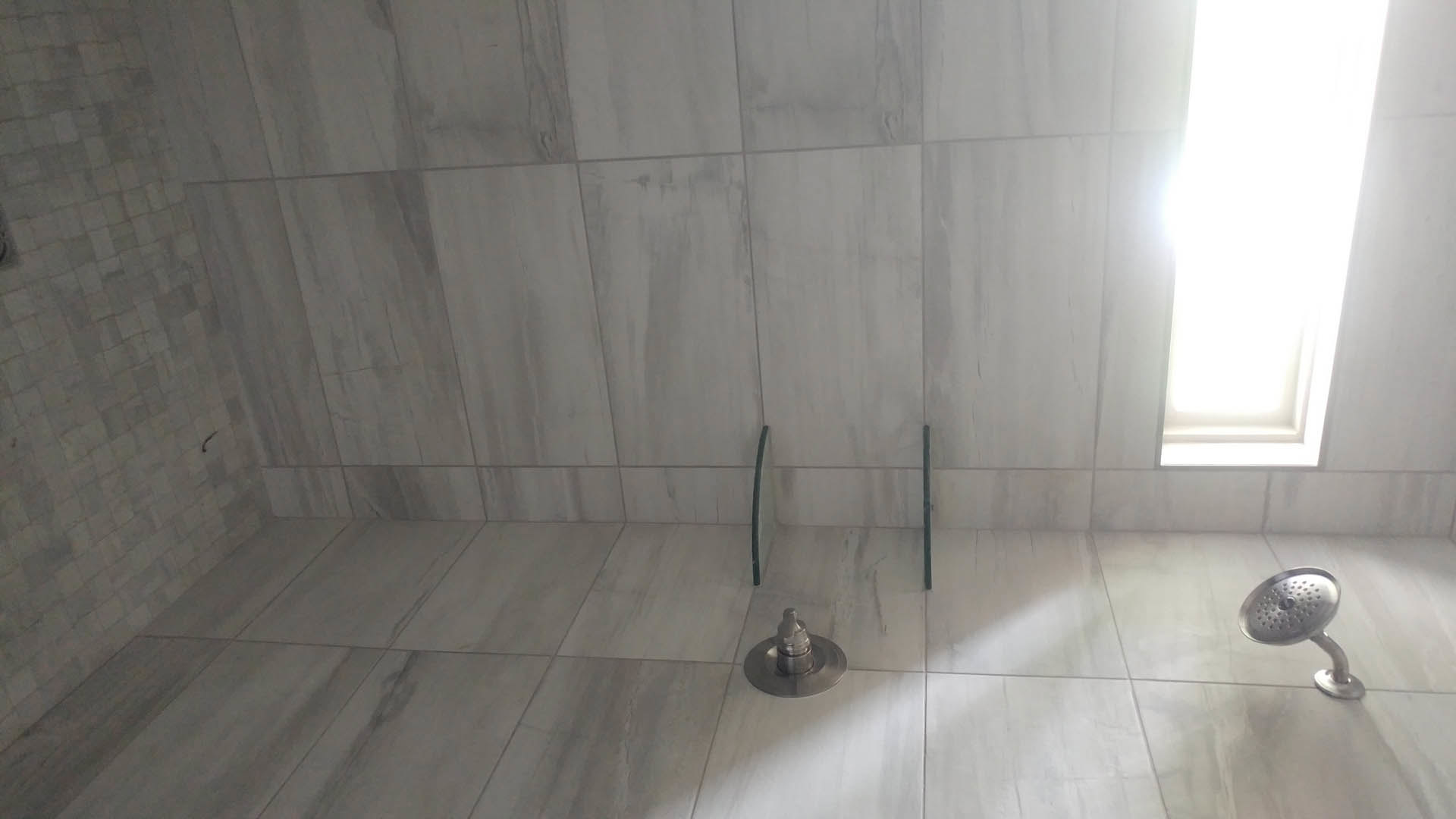 Circular metal shower head mounted on grey tiled wall, floor drain visible below, natural light streaming through window onto plaster walls and tile flooring.