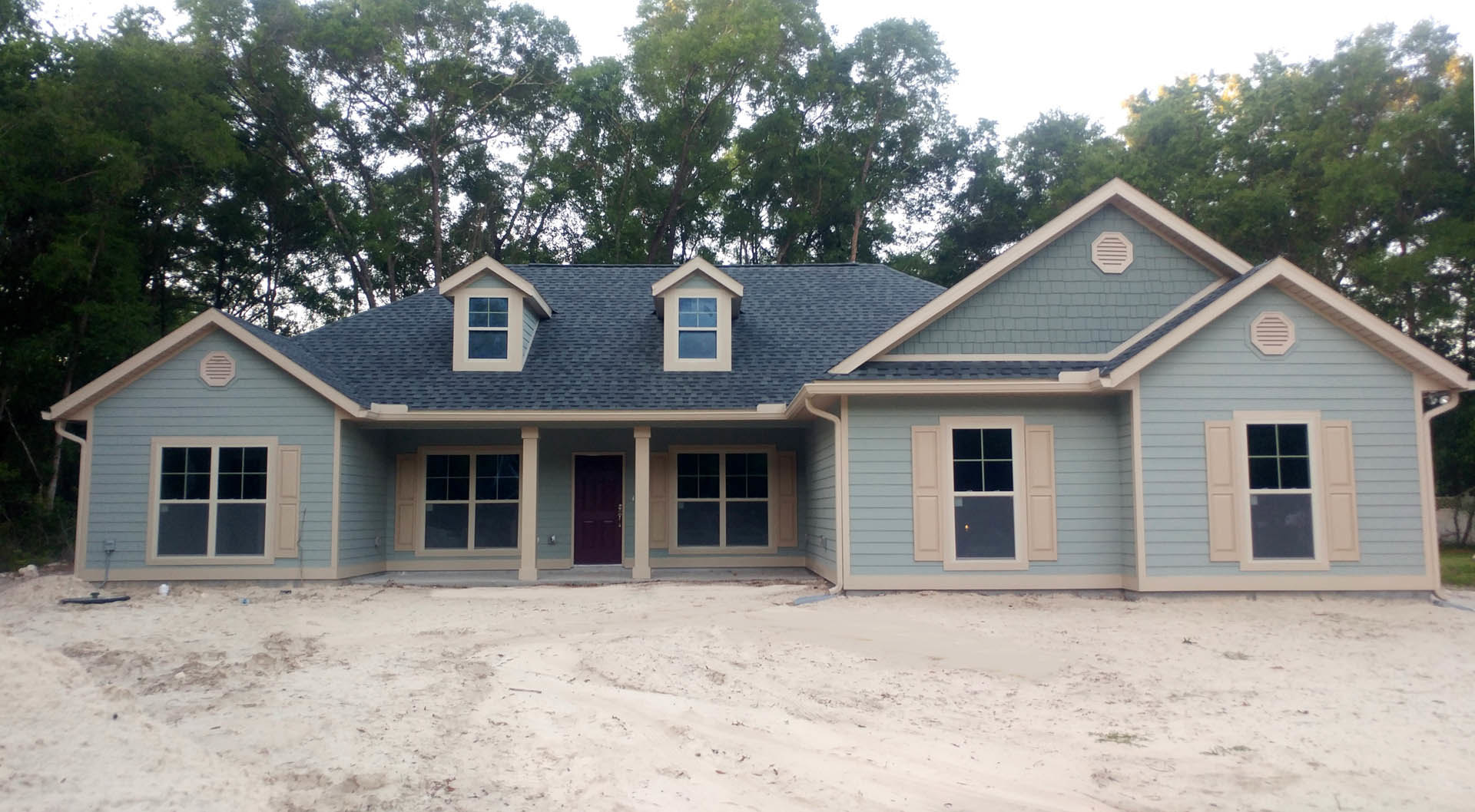 Two-story home with blue siding, white trim, red front door, and dirt driveway; mature trees and porch visible in front yard.
