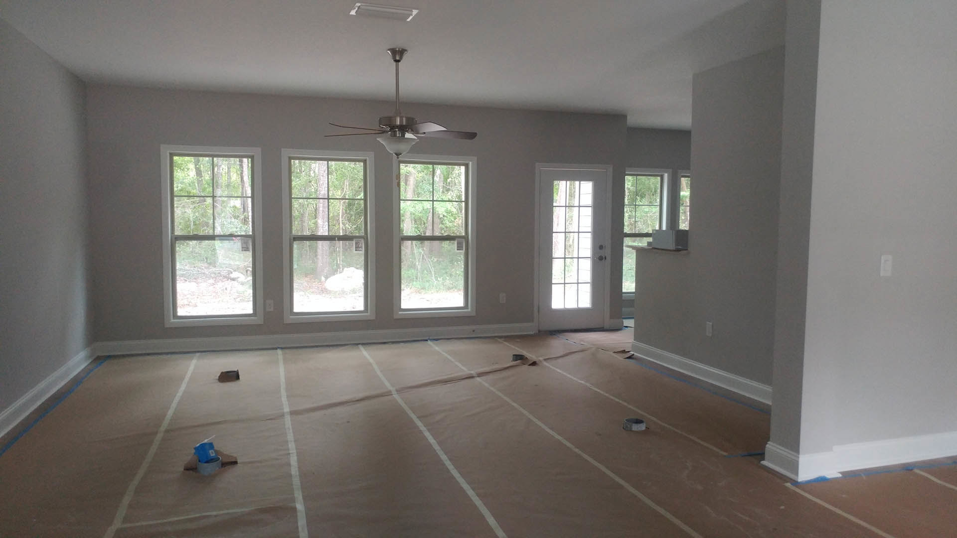 Living room with laminate wood flooring featuring white lines, ceiling fan with light, large windows showing trees outside, white door with glass panes, and a blue cup on a table.