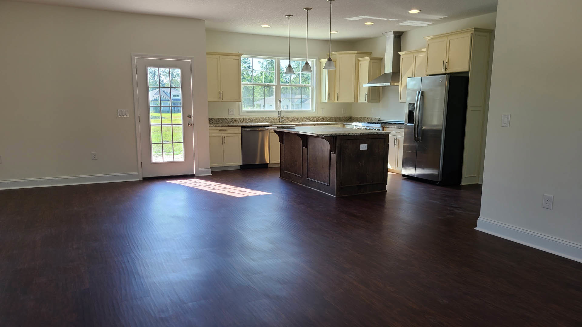 Kitchen with dark wood flooring, marble-topped island, stainless steel refrigerator, white cabinetry, and a door leading to a view of a neighboring house and trees through windows.