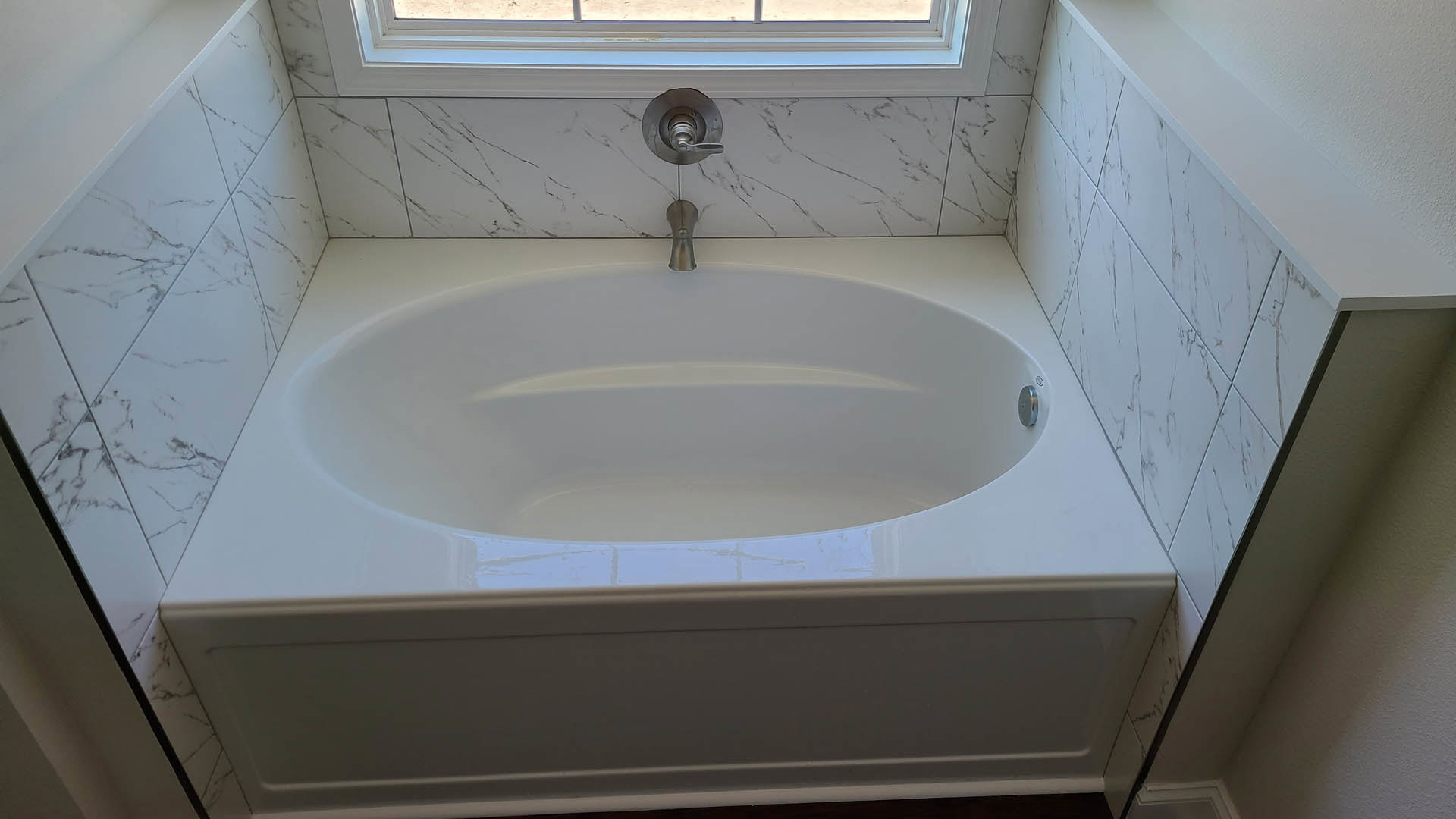 Freestanding white bathtub with chrome faucet beside a tiled wall and window in modern bathroom