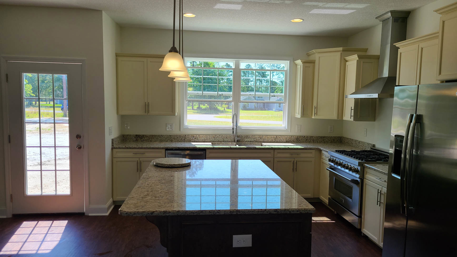 Spacious kitchen featuring a large marble island countertop reflecting natural light from a nearby window, stainless steel refrigerator with open door, white cabinetry, farmhouse