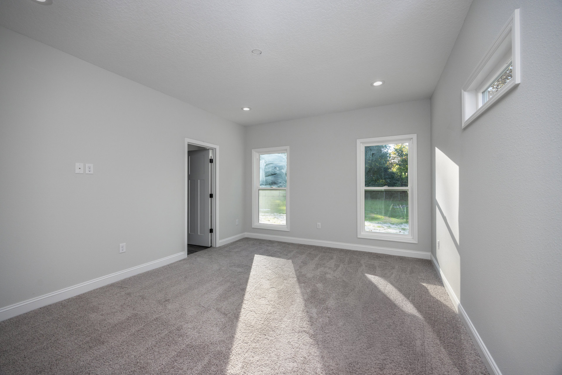 Carpeted room with white walls, large windows showing grass and trees outside, white door with black hinges, sunlight streaming in