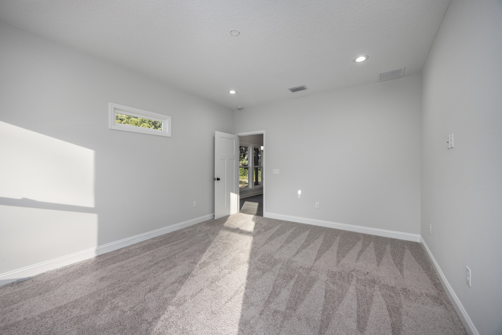 Open white door leading to a bright room with light wood flooring, smooth plaster walls, ceiling vent, and large window framing leafy trees outside