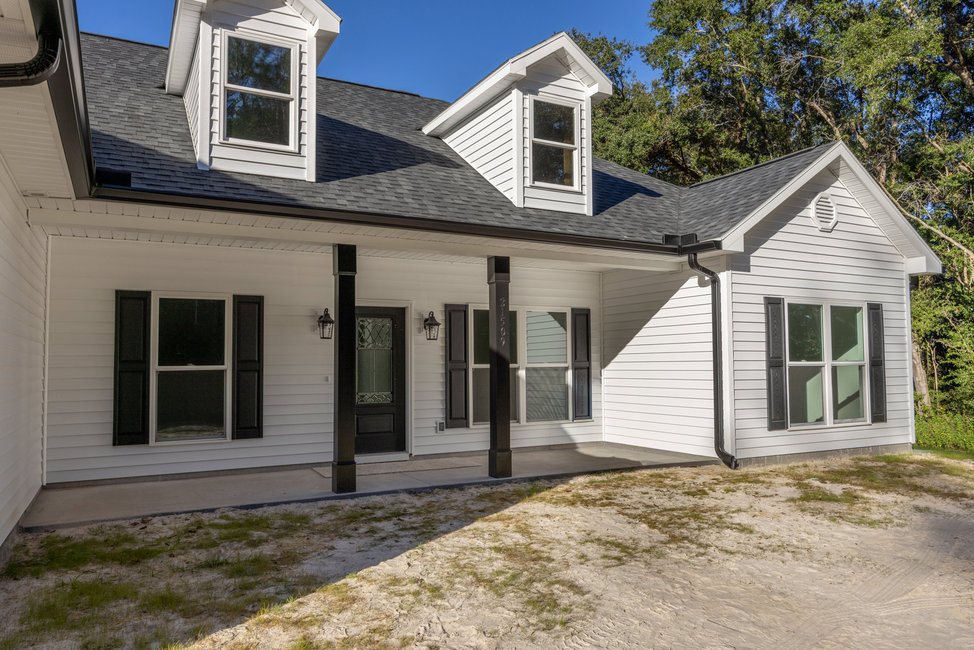 White siding house with black roof, black window shutters, black porch post, white entry door, and windows reflecting nearby trees