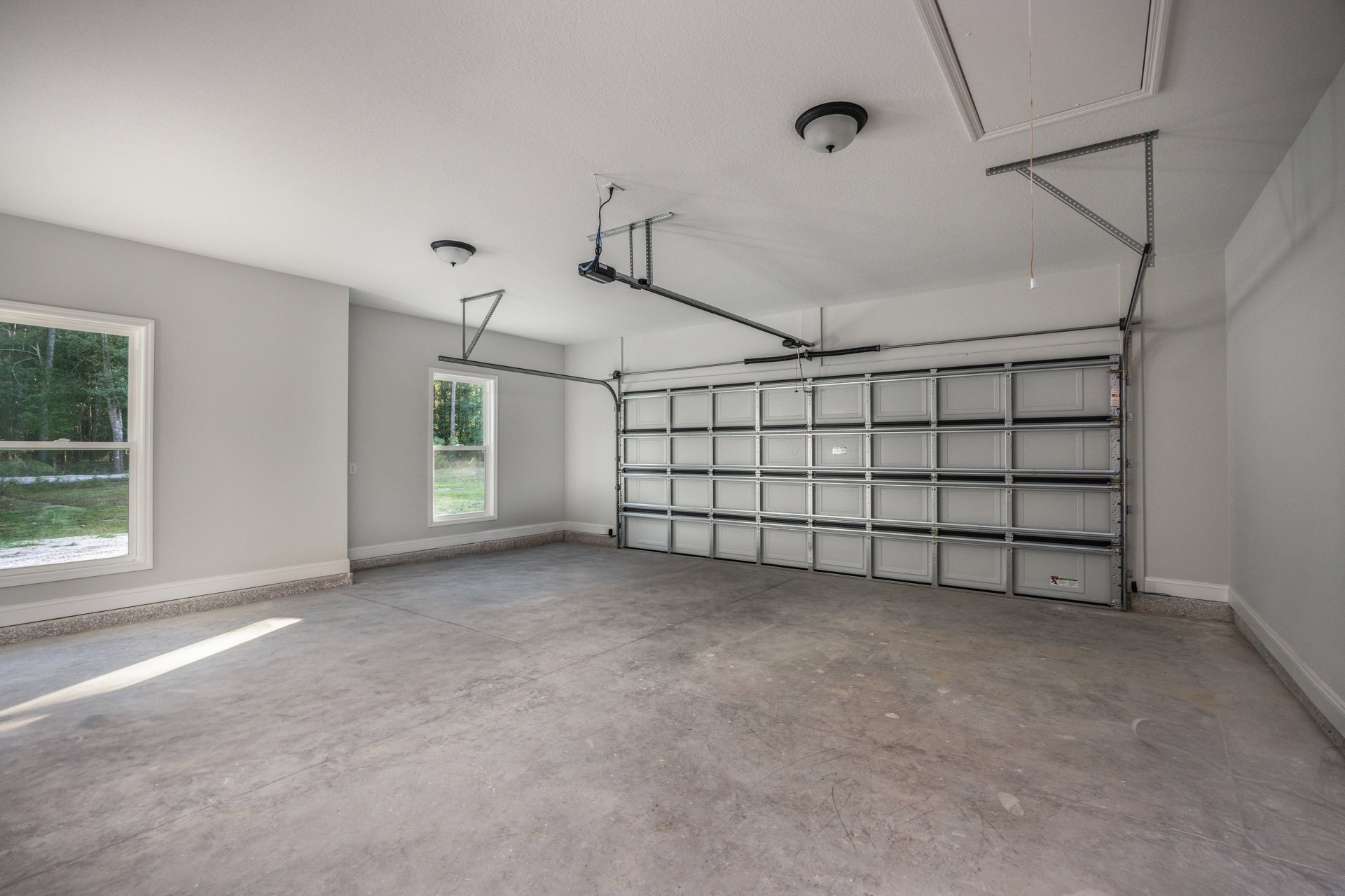 Spacious garage with smooth white ceiling, overhead light fixture, metal garage door, window overlooking trees, bare concrete floor