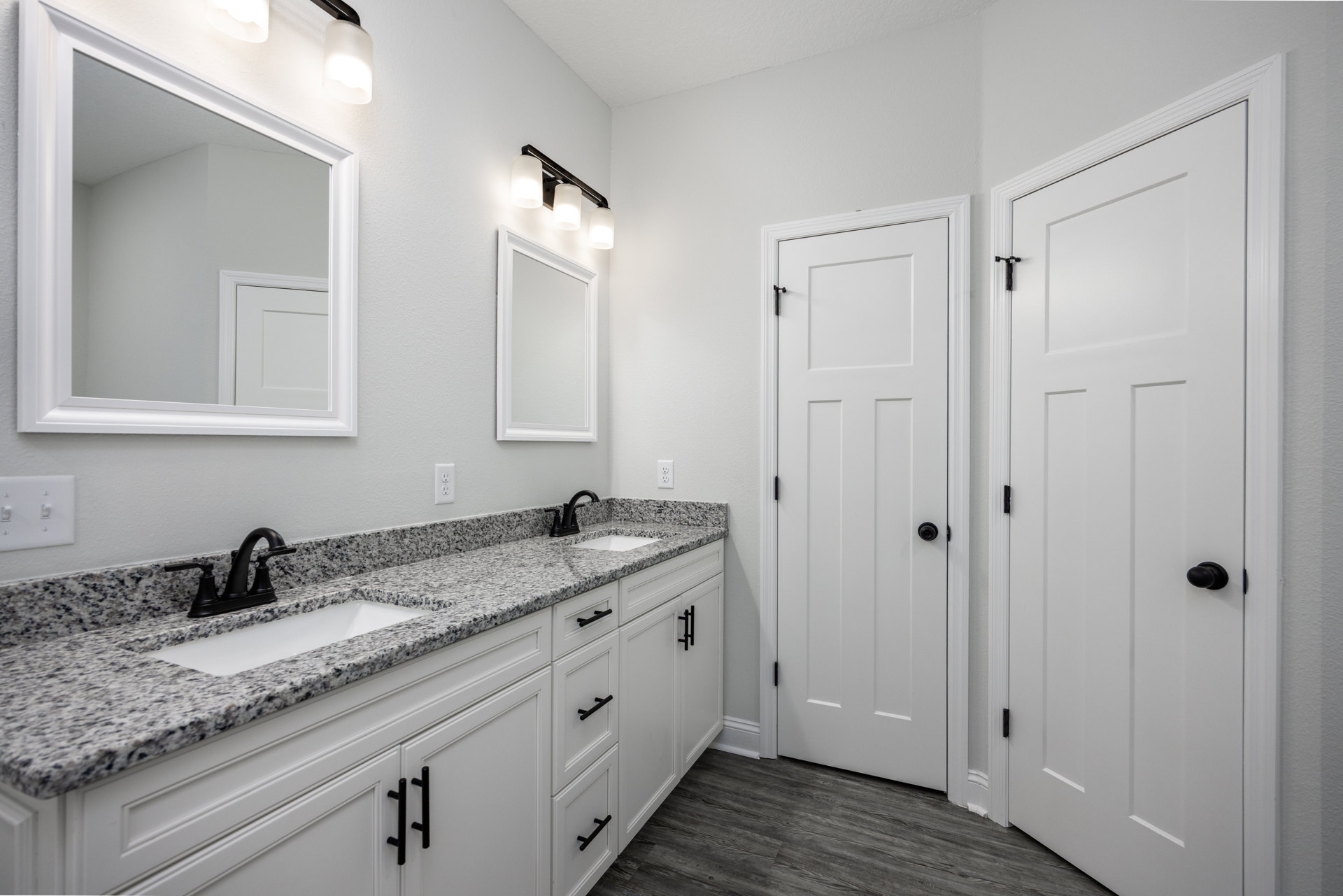 Bathroom with double sinks, black faucets on marble countertop, white rectangular mirrors above, white door with black knob, light tile flooring, white cabinetry