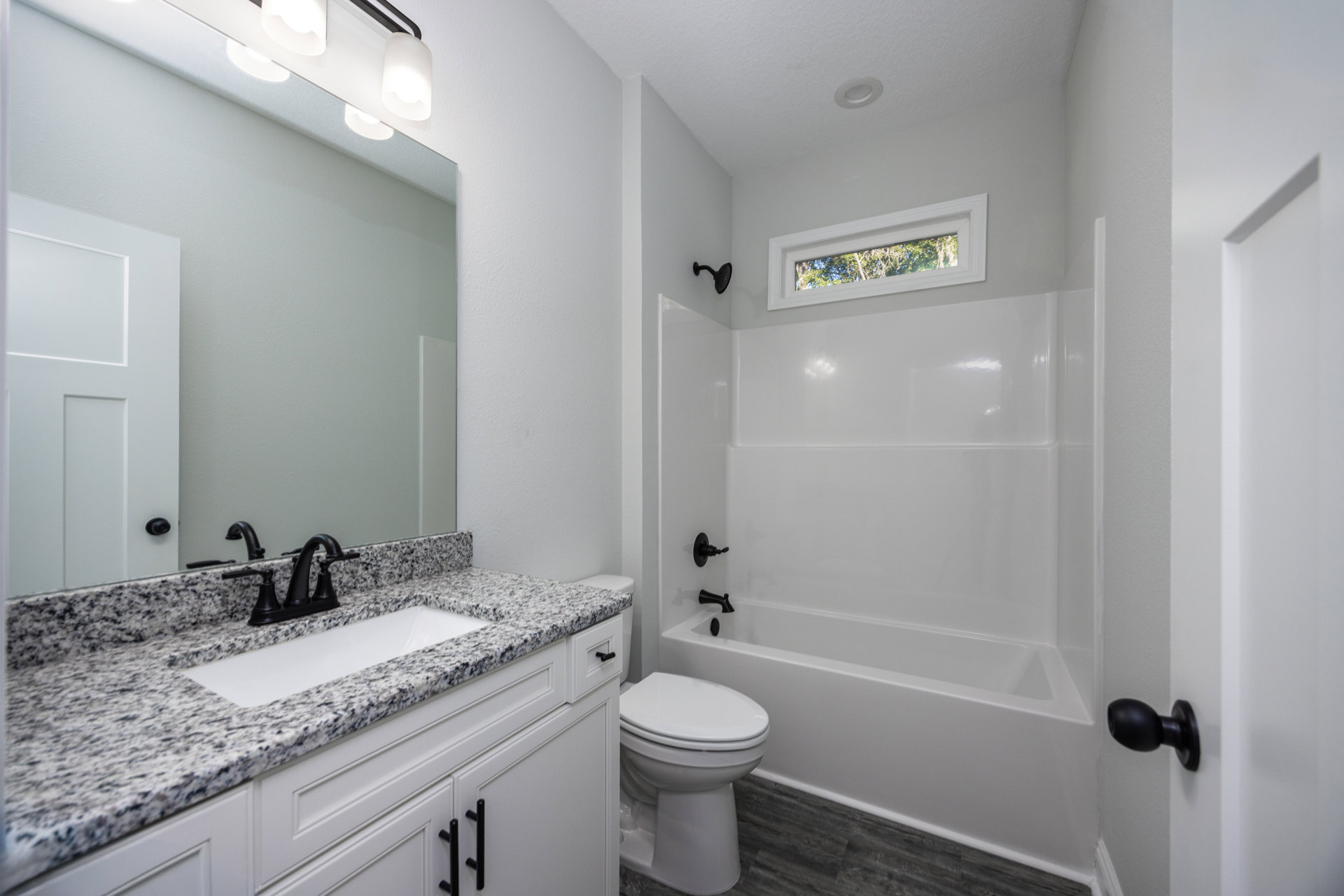 White porcelain sink with chrome faucet set in a light countertop, adjacent to a closed white toilet; large window reveals green trees outside, black towel hook mounted on white