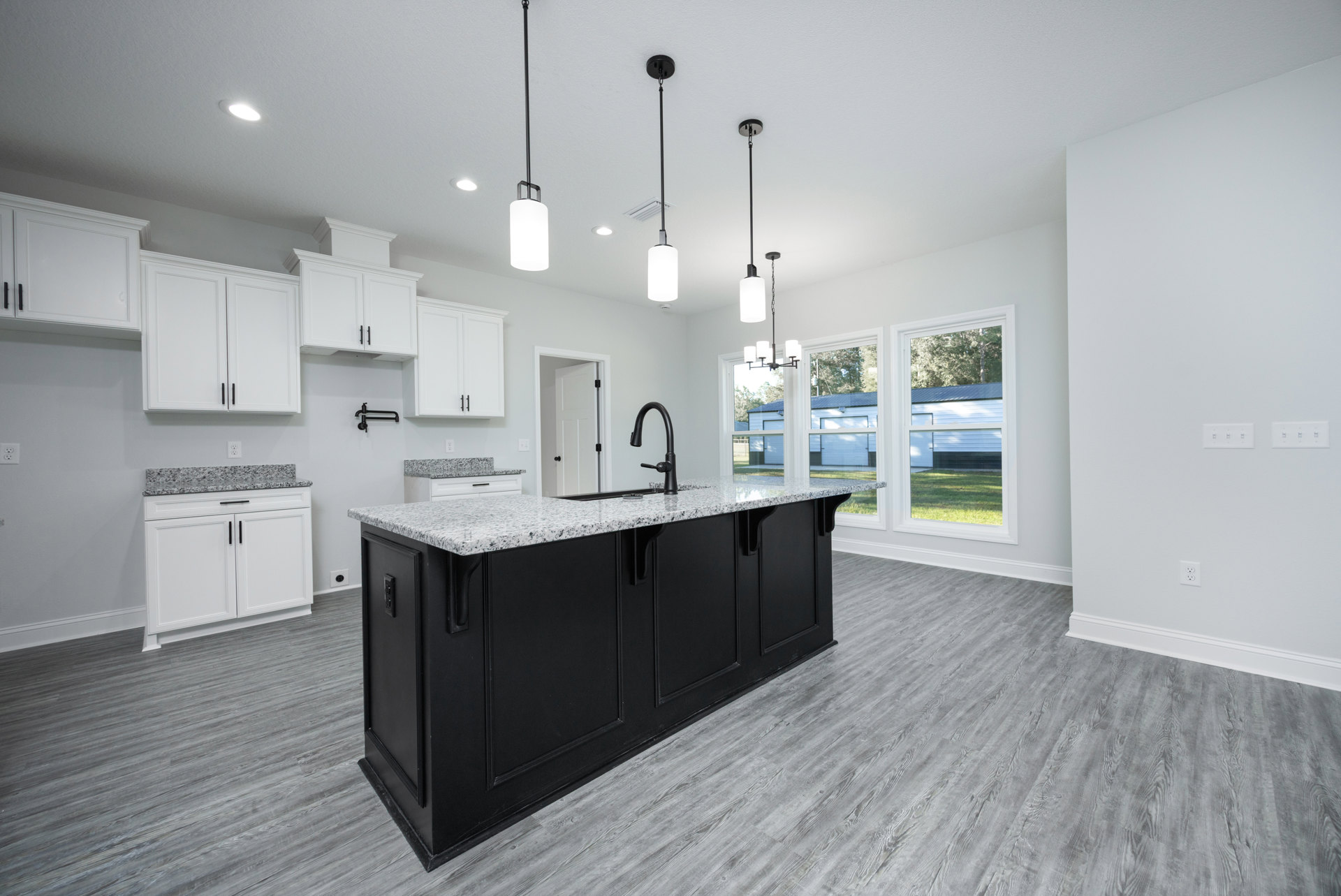 Black kitchen island with white countertop, surrounded by white cabinets with black handles, stainless steel sink, light fixture with black base, hardwood flooring, and neutral