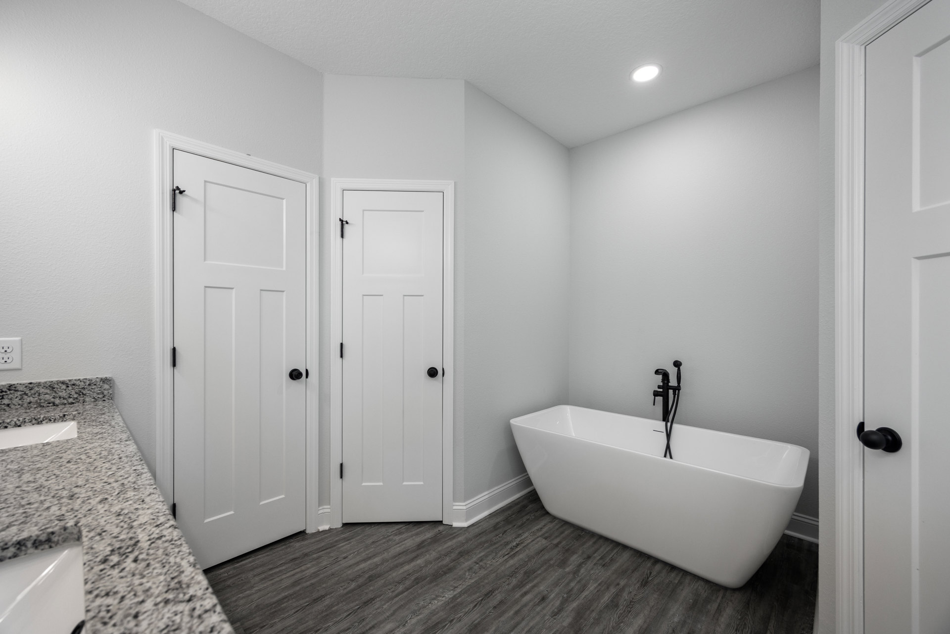 Bathroom with freestanding white tub, black faucet, double sink vanity with white countertop, two white doors with black hardware, light tile flooring and walls