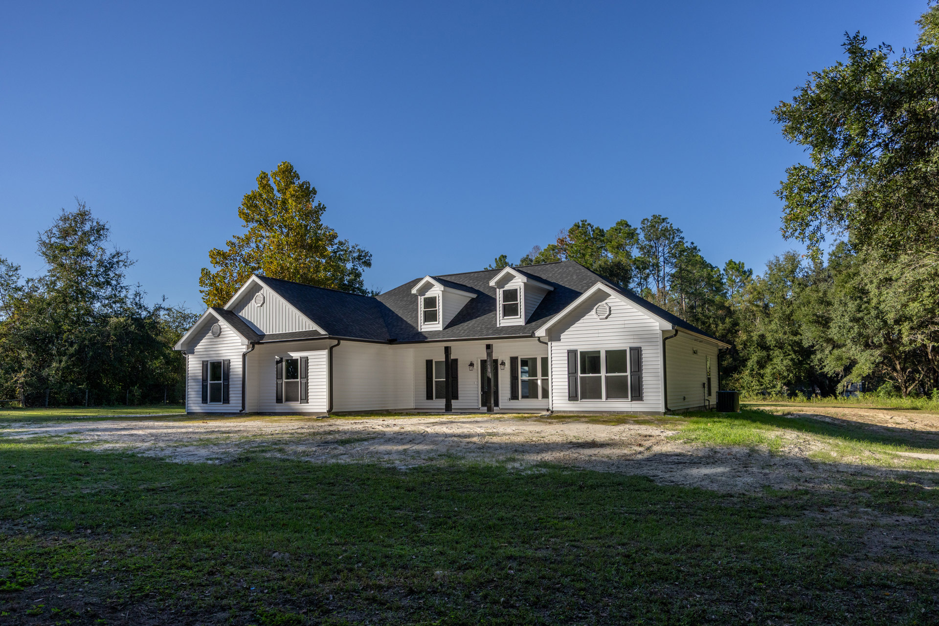 White farmhouse with black roof and shutters, green lawn in front, mature trees in background, broken window glass visible