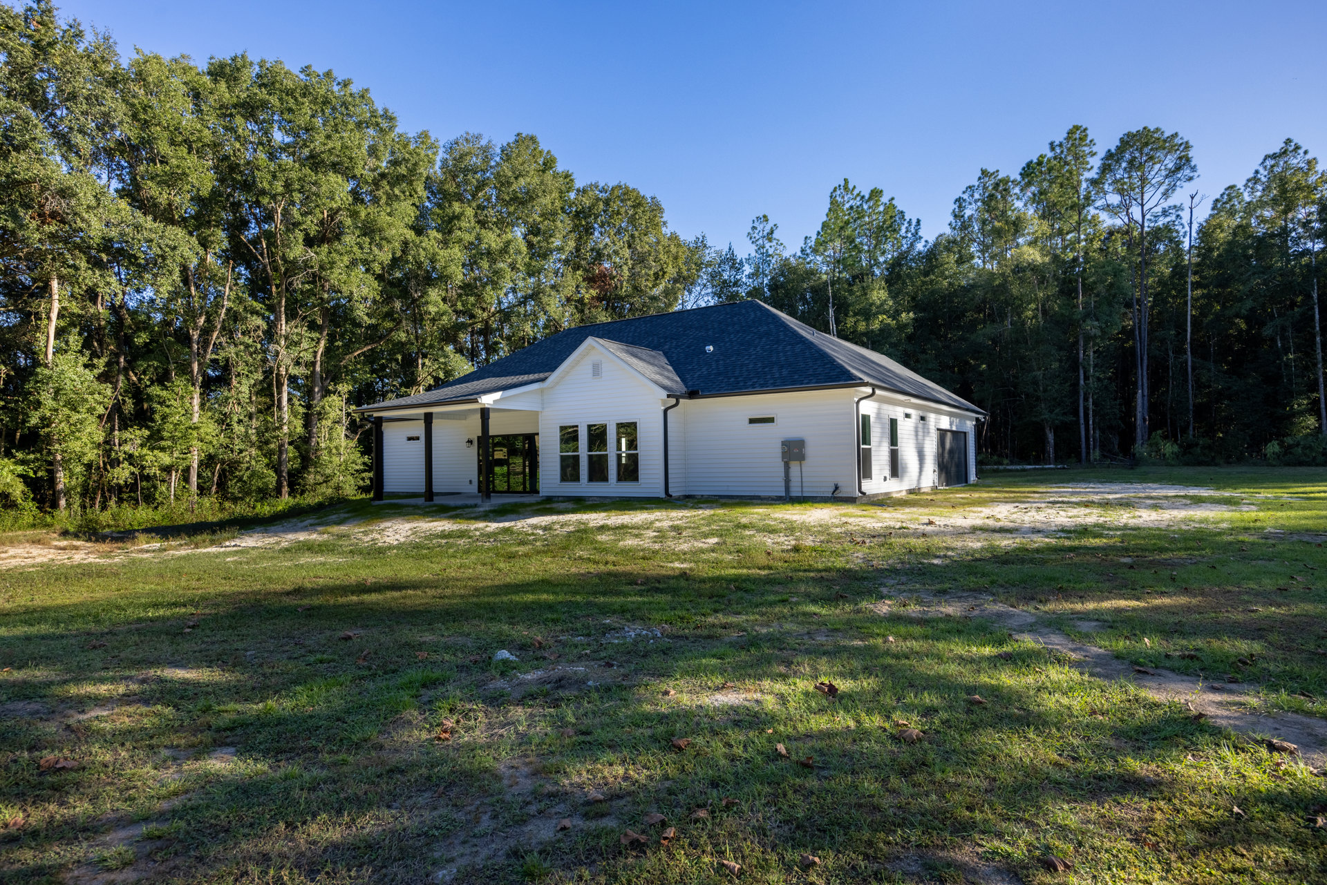 White house with black roof, attached garage, front lawn with scattered leaves, mature trees in background, multiple windows along facade