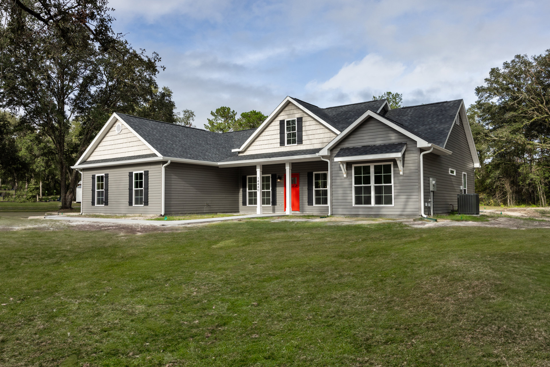 Two-story house with gray siding, red front door, white-framed windows, black heat pump unit, and green lawn under partly cloudy sky