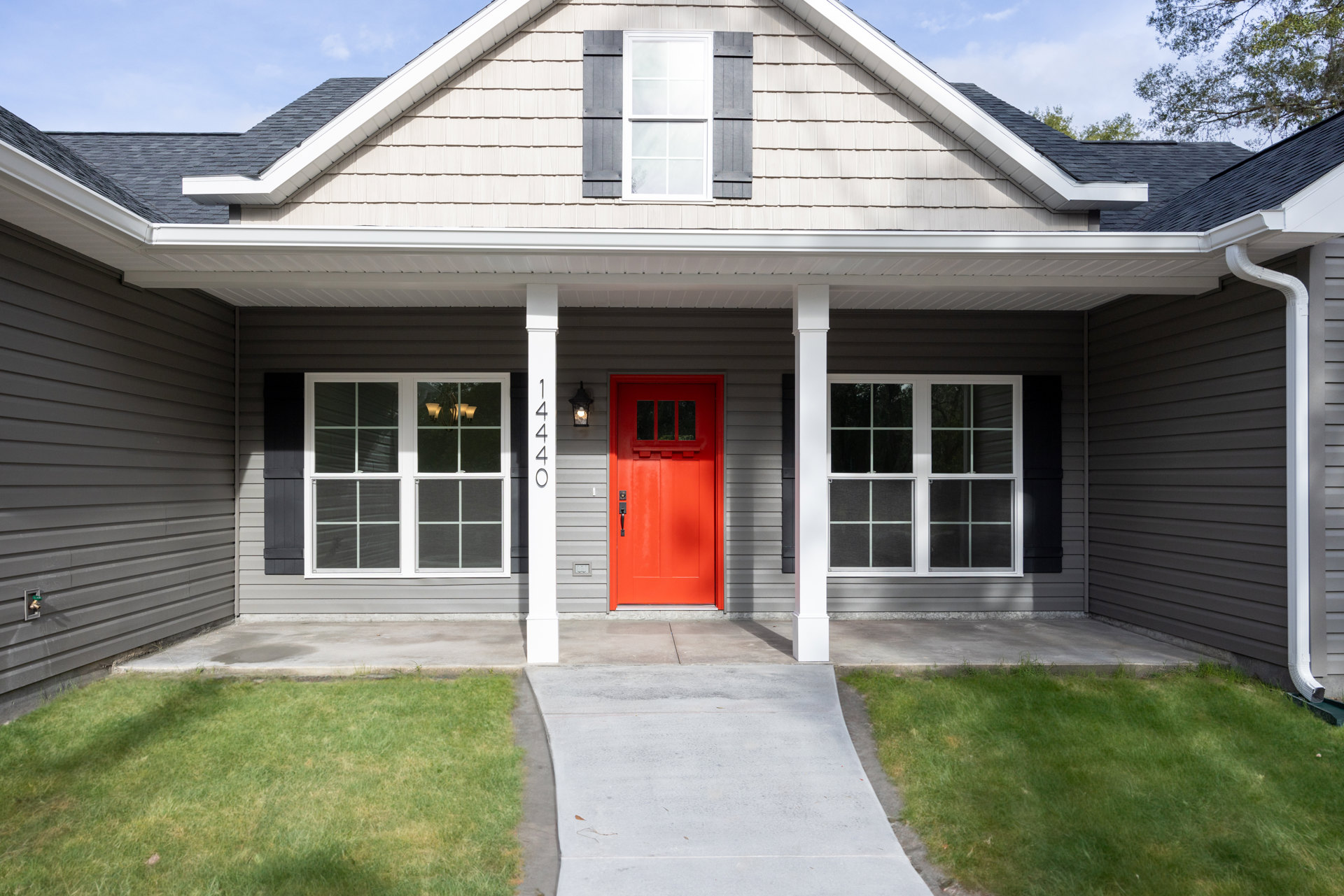 Red front door with black handle, white grid windows, light fixture visible inside, gray concrete walkway and ramp, light siding, green lawn, blue sky