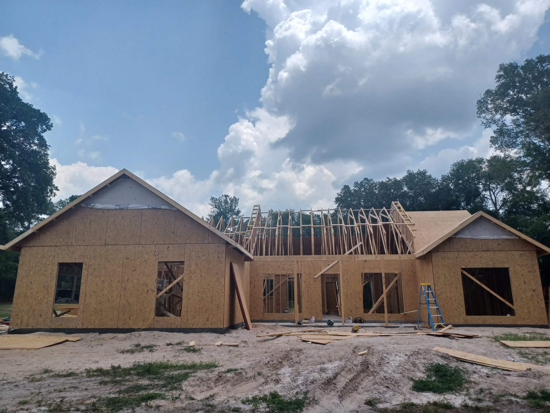 Framed house under construction with exposed wooden beams, partially completed roof, sandy ground with lumber pile, blue ladder inside, leafy trees in background, cloudy sky