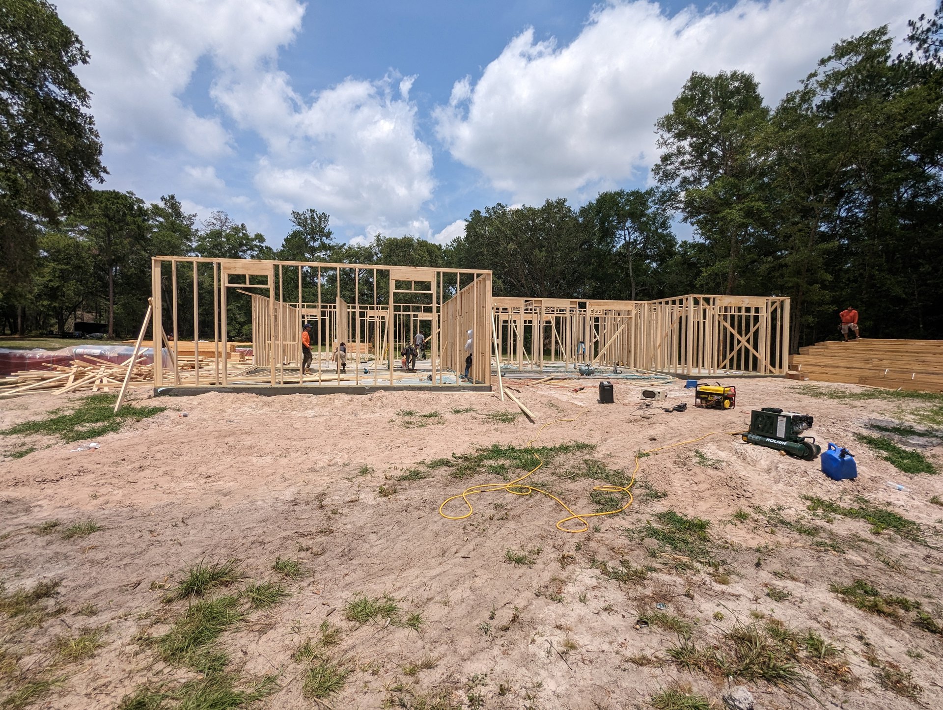 Wood-framed house under construction with exposed beams, workers on site, blue plastic canister and construction equipment on dirt ground, trees and partly cloudy sky in background