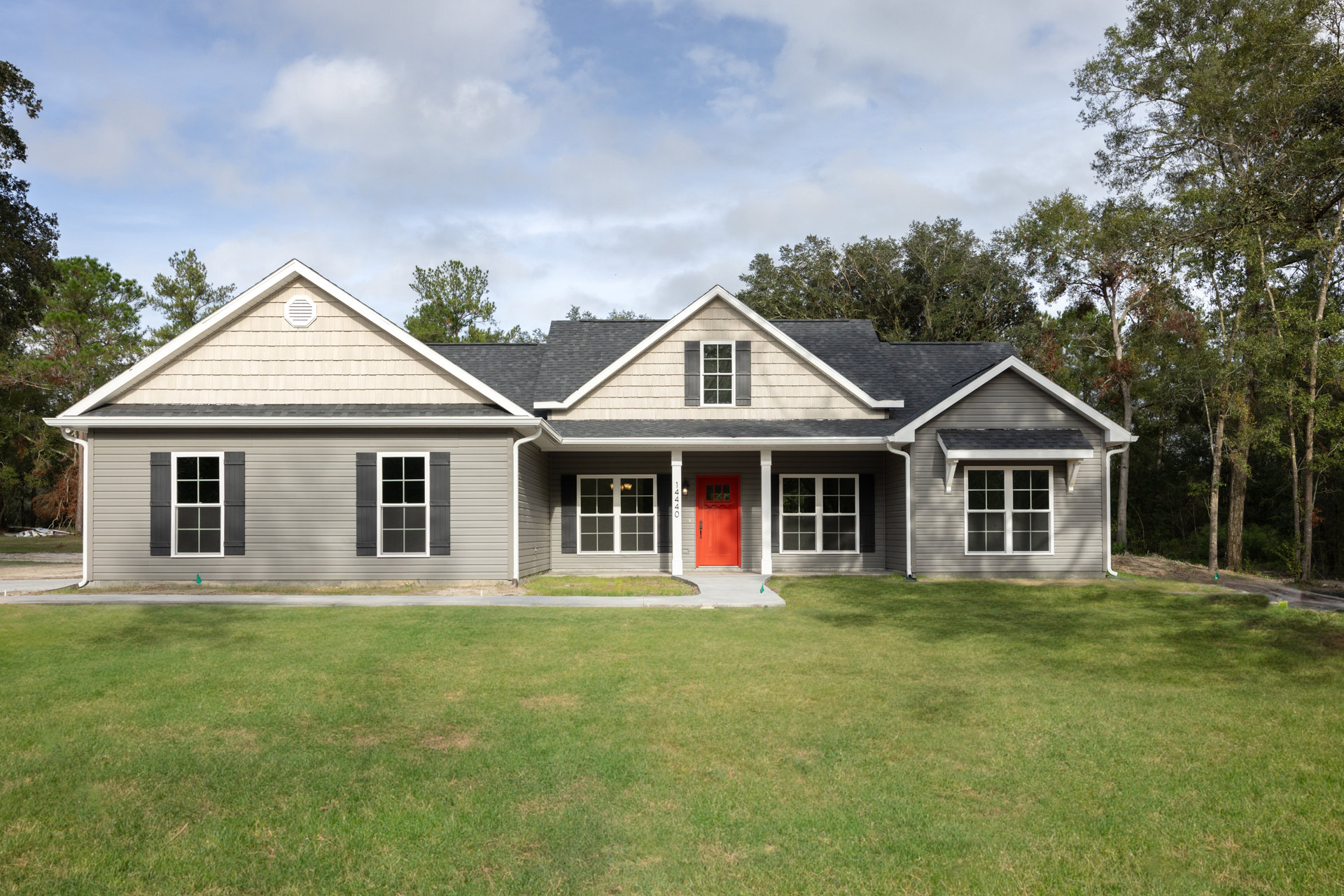 Two-story home with gray siding, white trim, and a prominent red front door; covered porch with white columns, multiple windows with white frames, manicured lawn, and cloudy sky