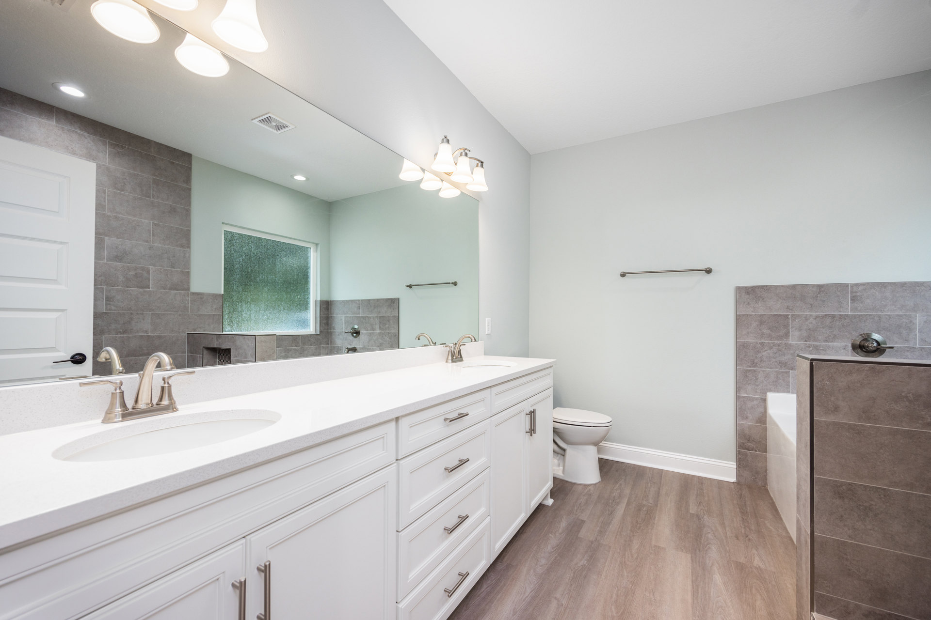 Bathroom with expansive mirror above white countertops, white toilet, glass window with white frame, modern light fixture, white door, tiled floor, and close-up of chrome handle.