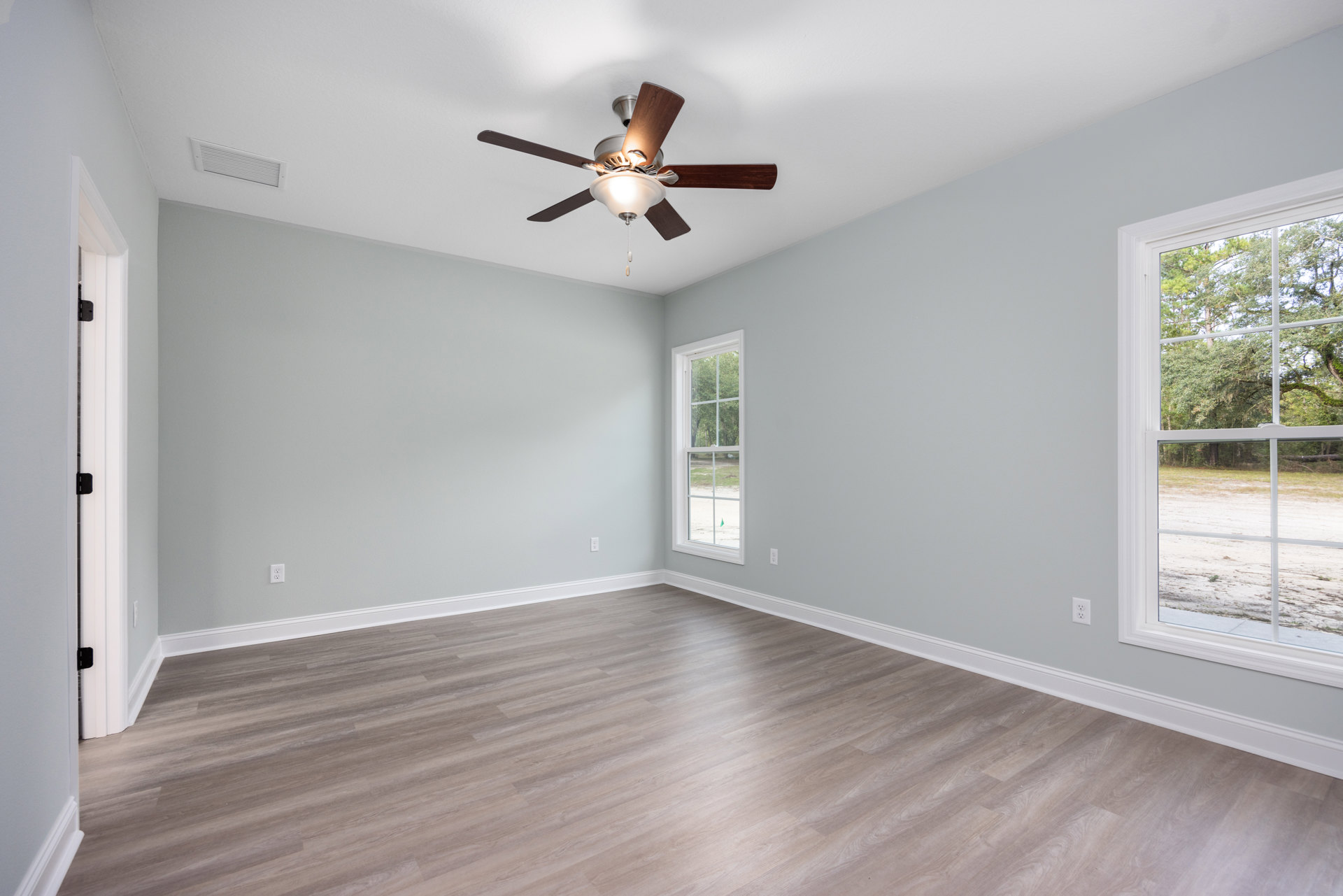 Ceiling fan with light fixture above hardwood floors, white plaster walls, large window overlooking grass, trees, and a dirt road