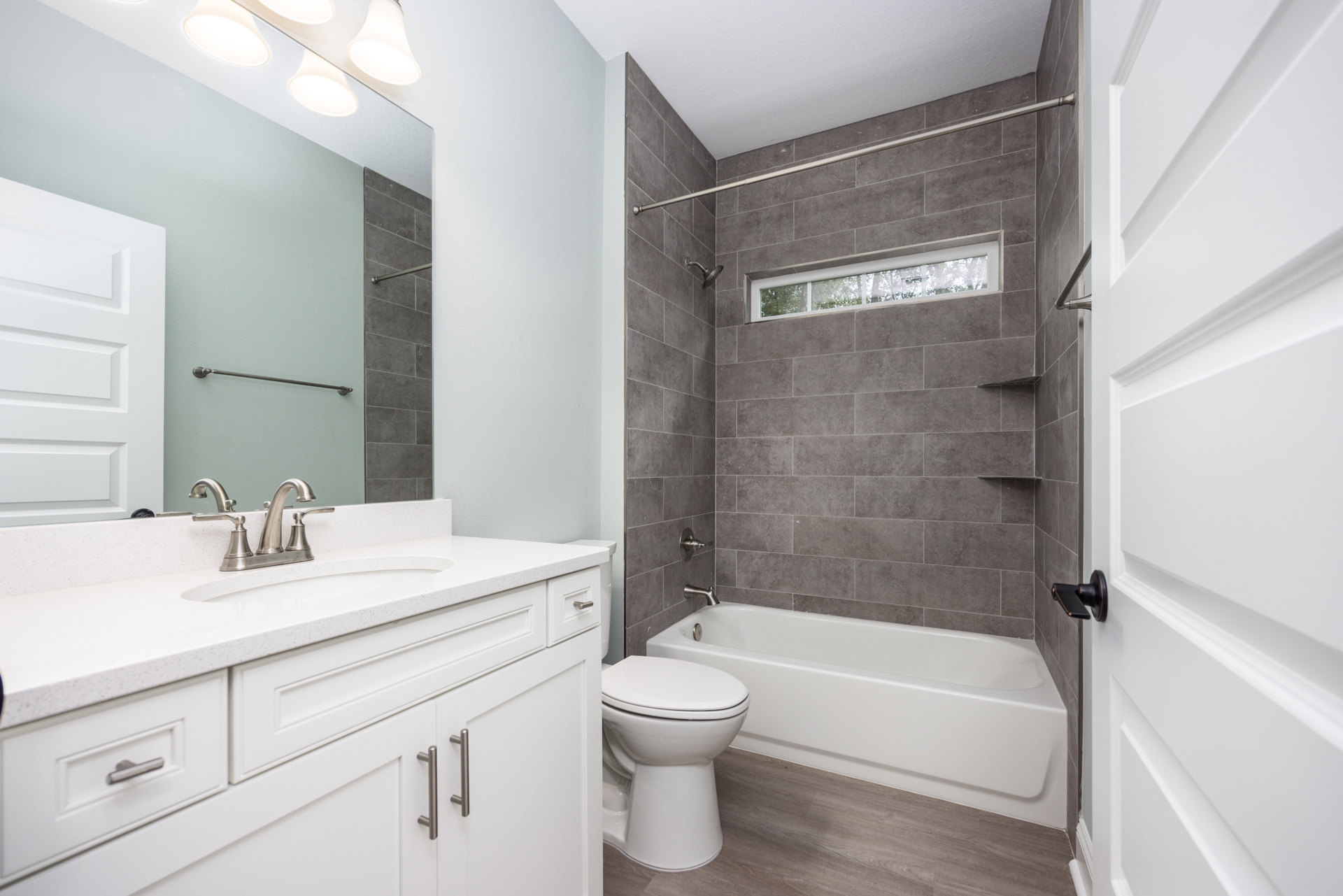White bathroom with tile floor, white toilet beside a built-in bathtub, white vanity with sink and chrome faucet, rectangular mirror above, white door with black handle.