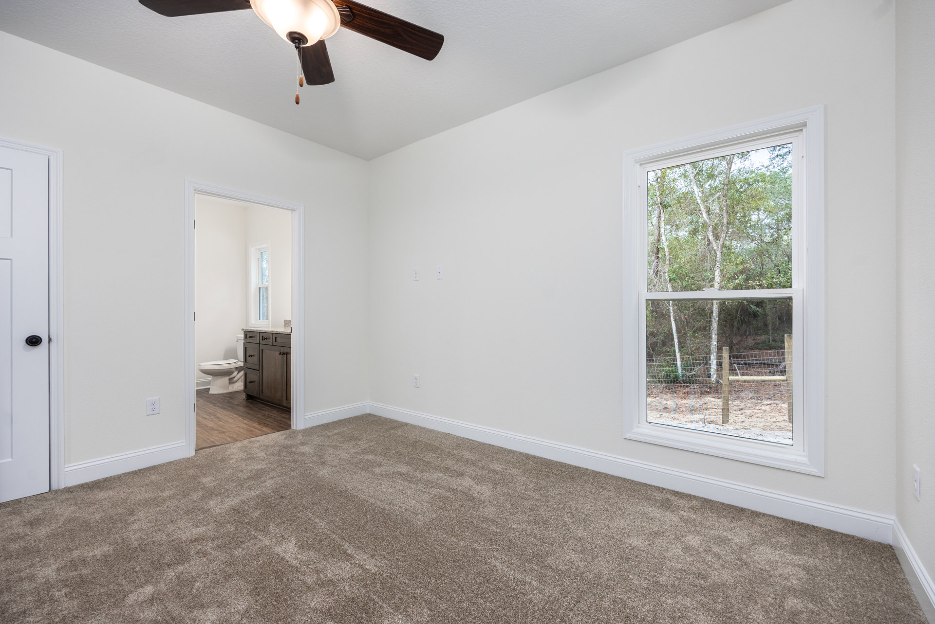 Bedroom with beige carpet, white walls, ceiling fan with light, single window showing green trees, white toilet partially visible, wood cabinet in corner