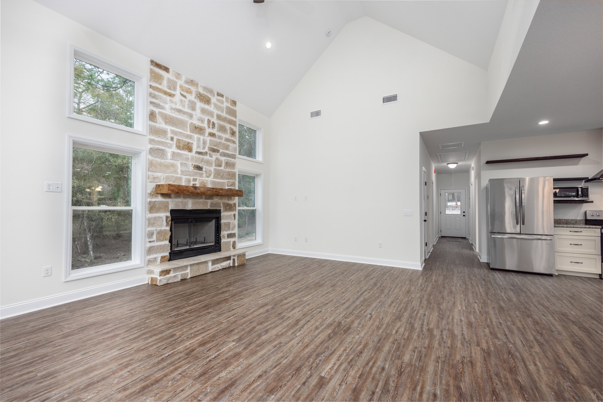 Living room with wood flooring, stone fireplace featuring a mesh screen, stainless steel double-door refrigerator, large window showing trees outside, and a white door with glass