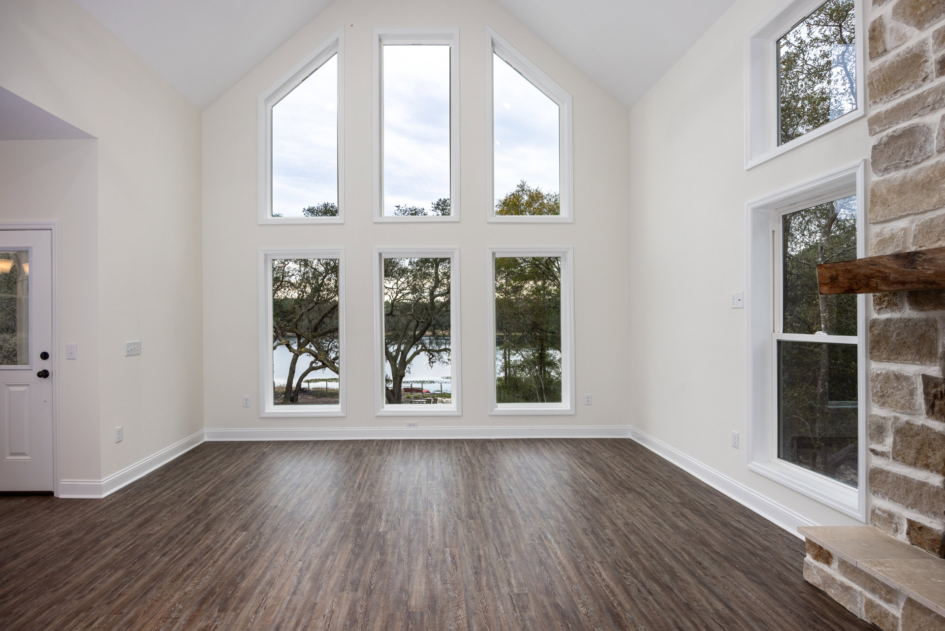 Spacious room featuring wood flooring, white walls, and multiple large windows overlooking moss-covered tree branches and a cloudy sky