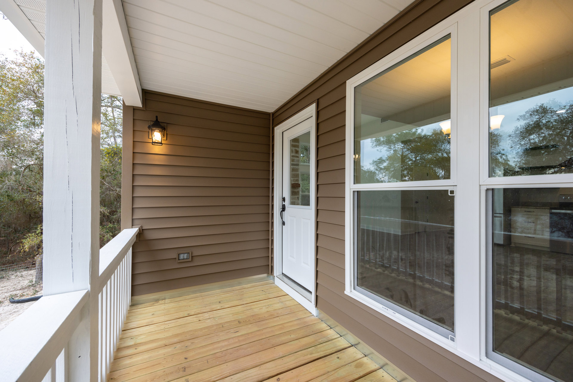 White front porch featuring a white door with black handle, adjacent window, wood flooring, white ceiling with brown paneling, and wall-mounted light fixture.